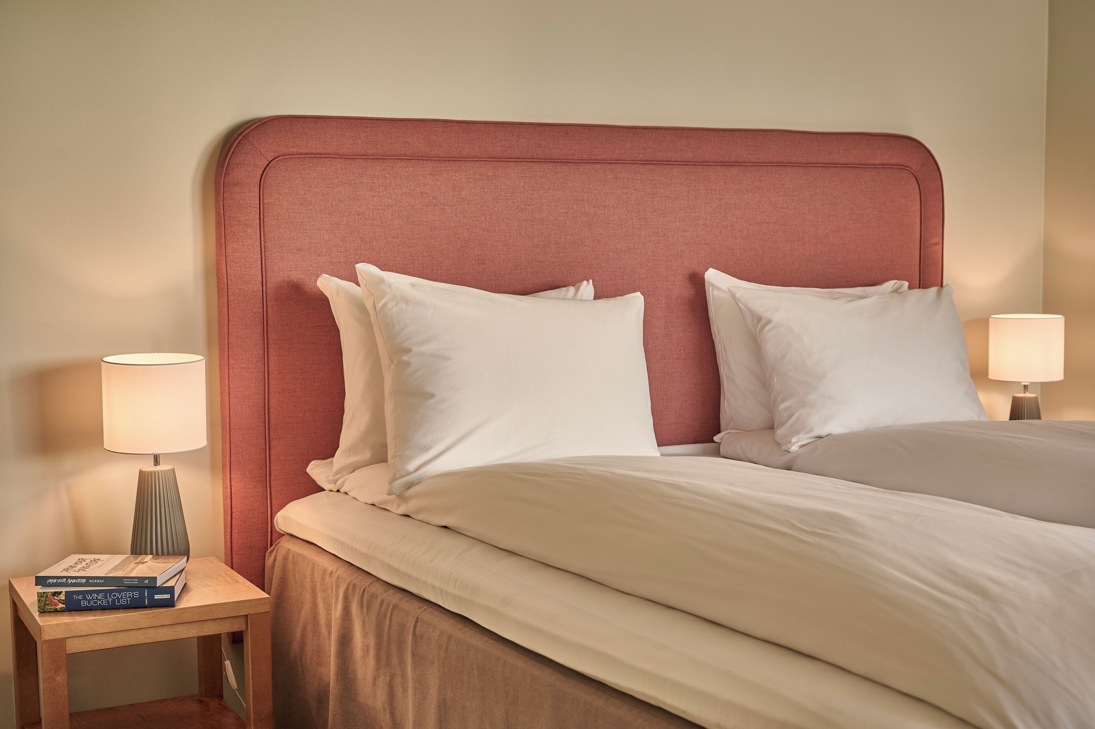 Quarto de hotel com uma cama grande, duas luminárias laterais, uma mesinha com livros e um painel de cabeceira vermelho.