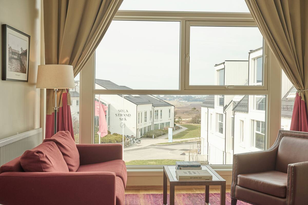 A room with a large window, a sofa, a chair, and a table with a book.