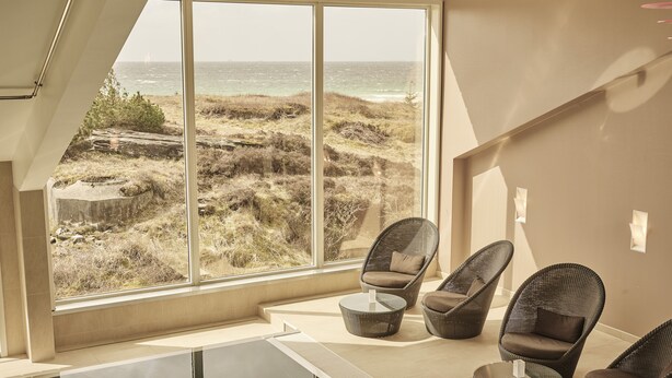 A modern interior with large windows overlooking a coastal landscape, wicker furniture, and a glass table. - Sola Strand Hotel (Sola)