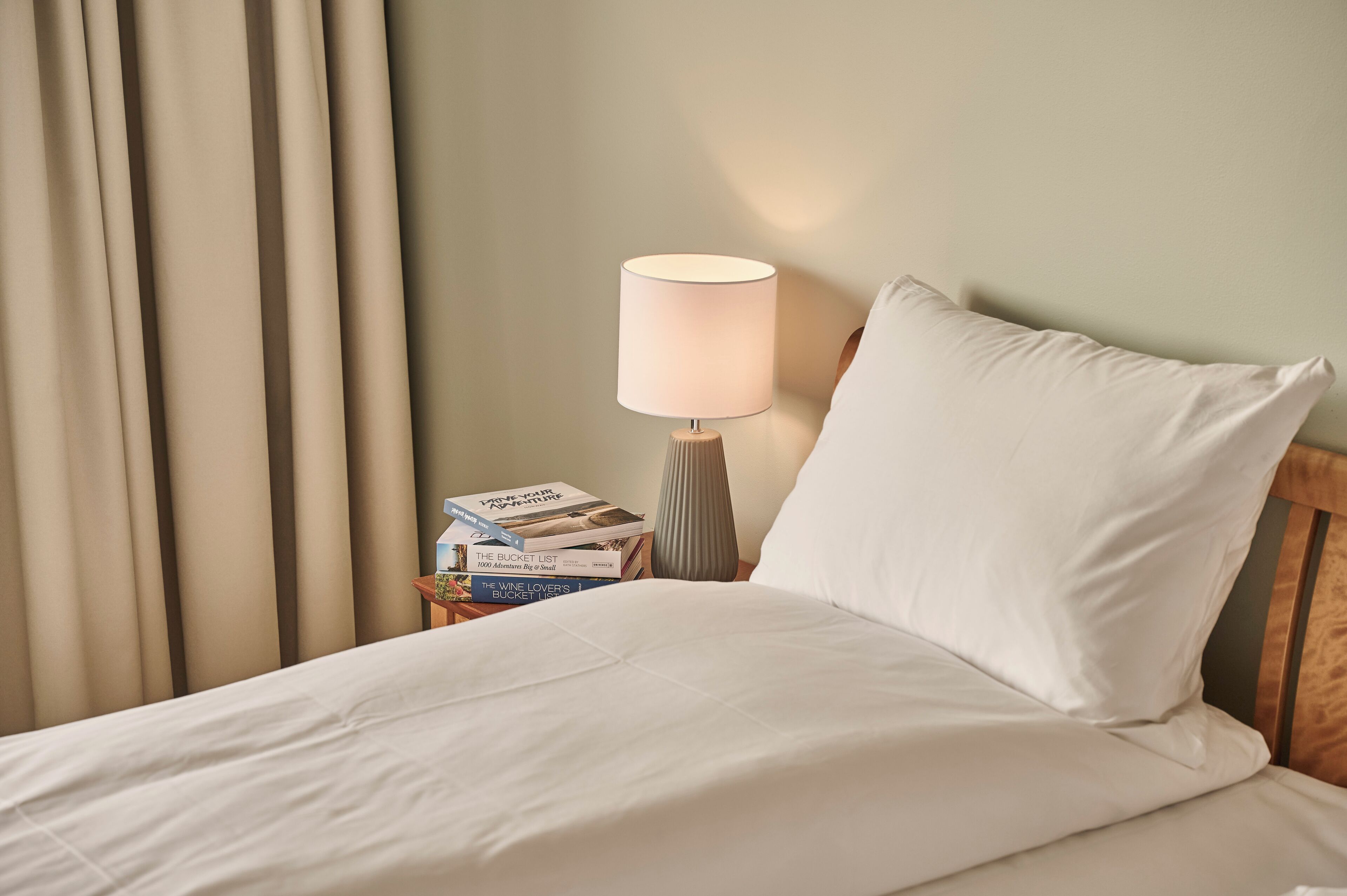 A neatly made bed with white linens, a bedside lamp, and a stack of books on a nightstand.