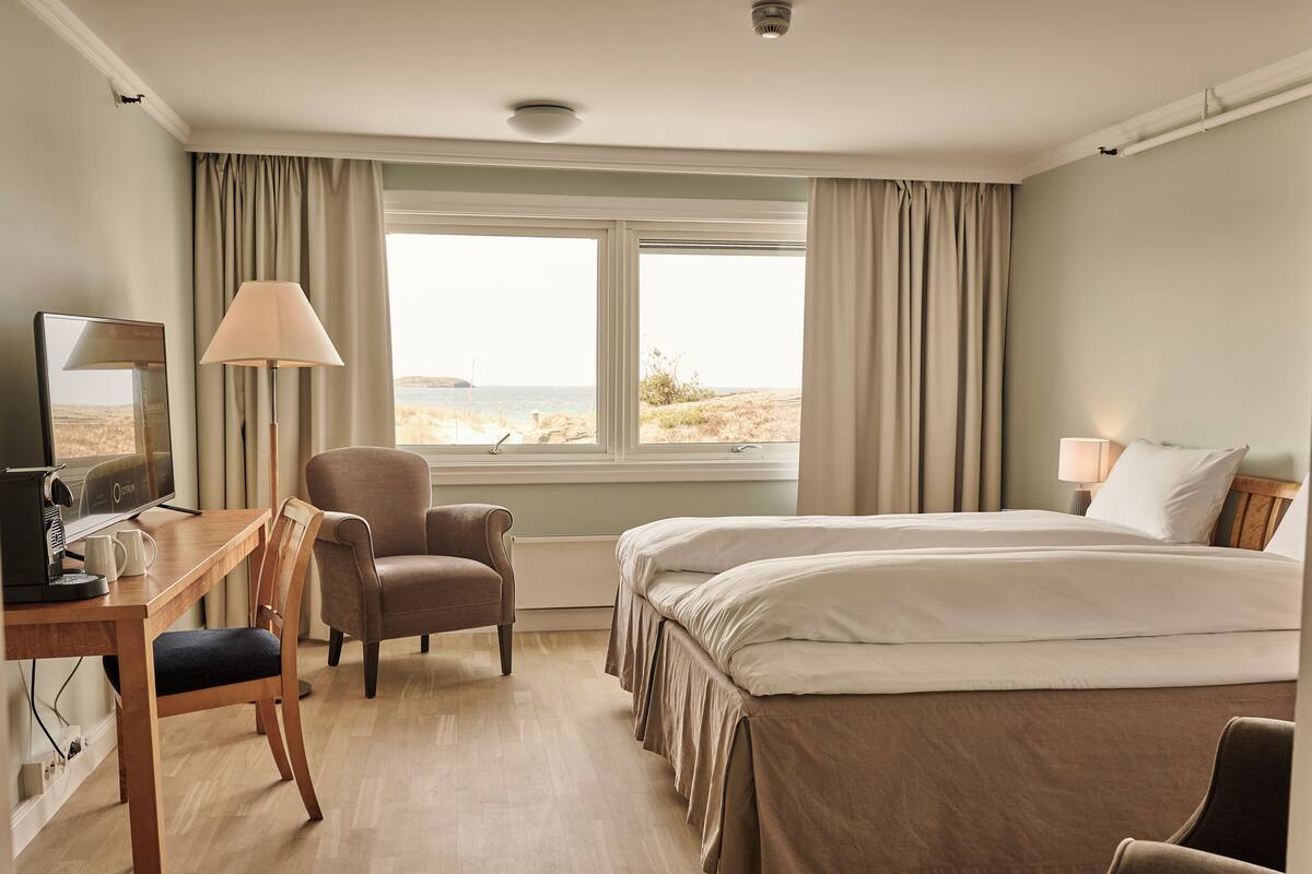 A hotel room with a bed, a desk with a computer and a lamp, a chair, and a view of the sea through the window.