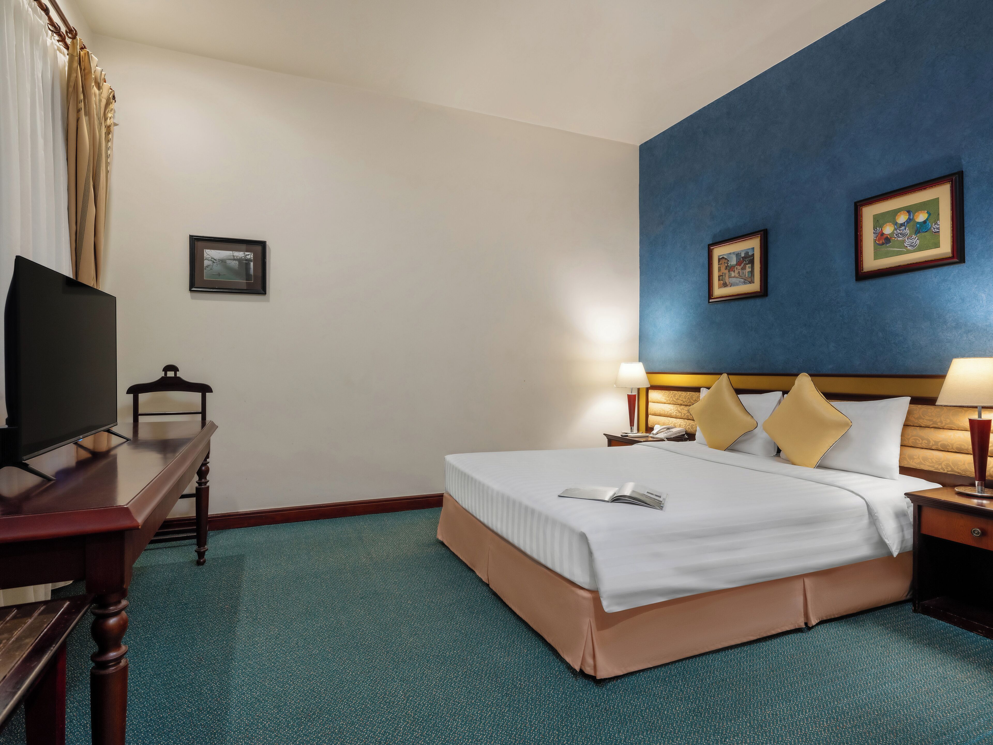 A hotel room with a bed, a desk, a television, and two paintings on the wall.