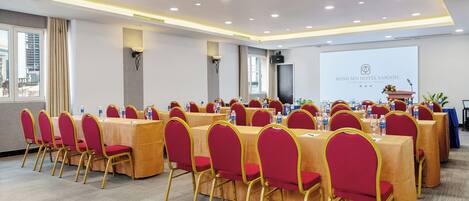 A conference room with multiple tables and chairs, equipped with water bottles and glasses, and a presentation screen in the background.