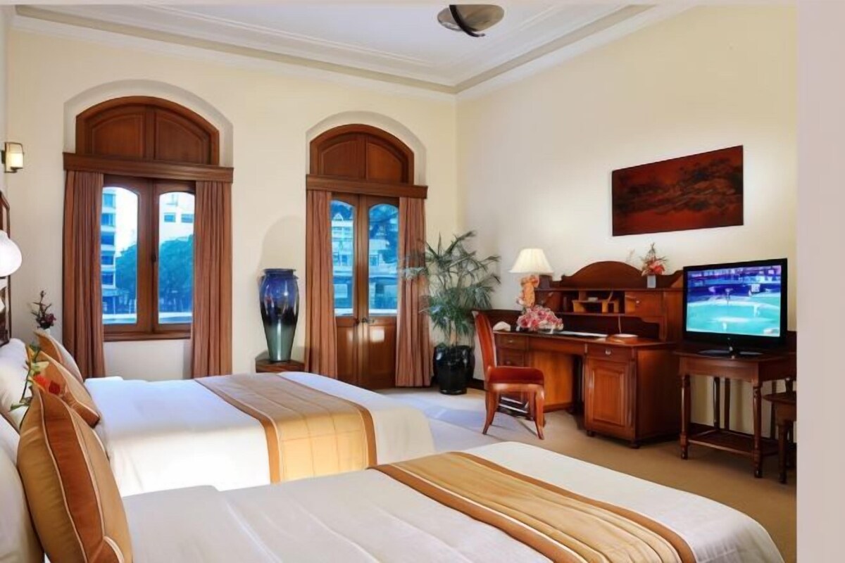 Une chambre d’hôtel avec deux lits, un bureau avec une télévision, une plante en pot et une fenêtre avec des rideaux.