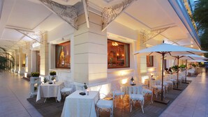 An outdoor dining area with white tables and chairs, covered by a large canopy, and illuminated by ambient lighting.
