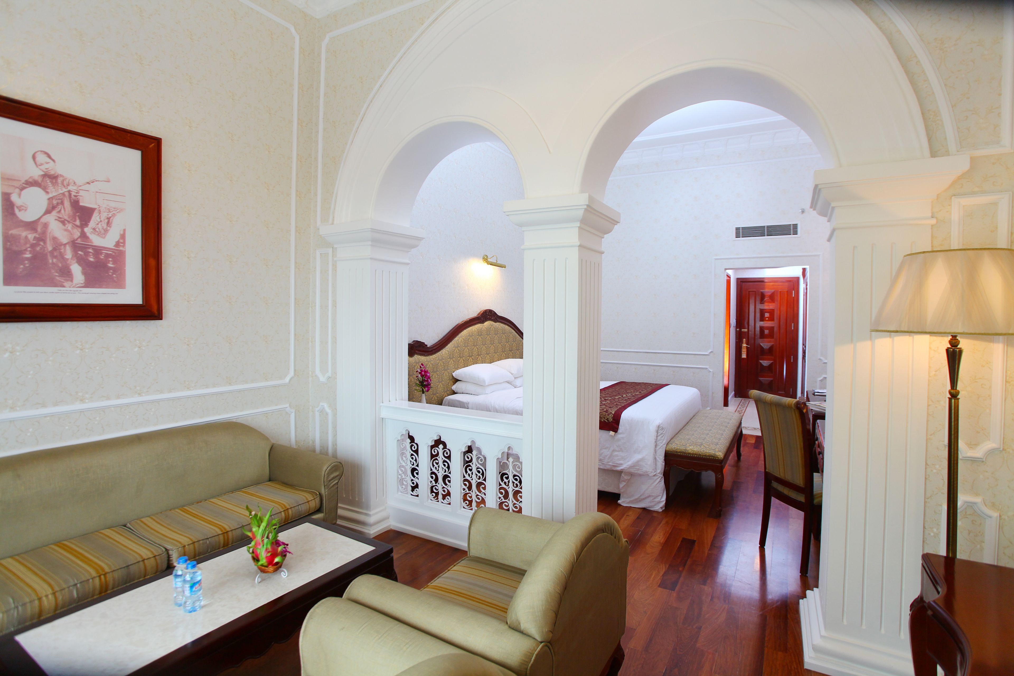 A hotel room with a sofa, a small table with a vase and bottles, a bed with a wooden headboard, and an arched doorway leading to another room.