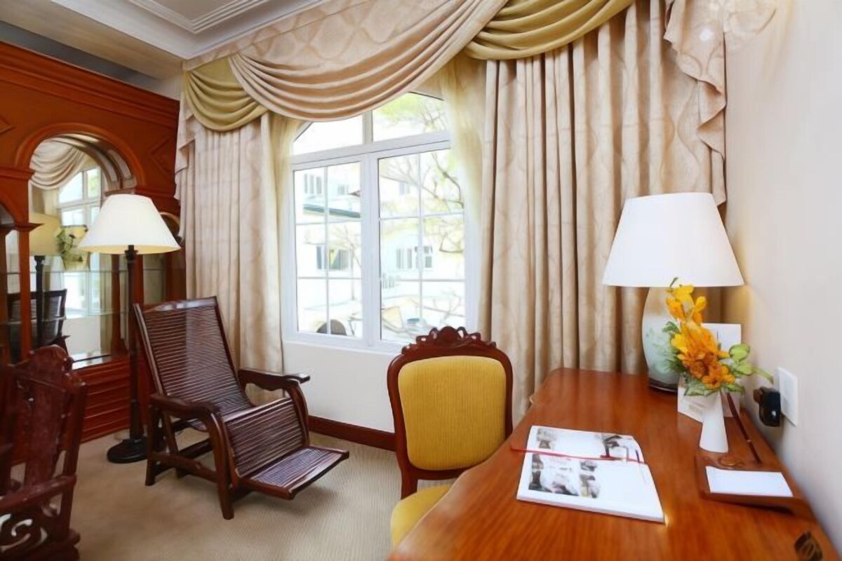 Une chambre d’hôtel avec un bureau en bois, deux chaises, une lampe et une fenêtre avec des rideaux.