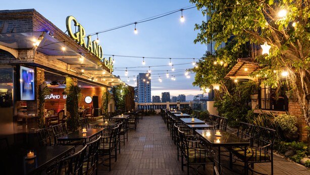 An outdoor dining area with tables and chairs, string lights, and a city skyline in the background. - Hotel Grand Saigon (Ho Chi Minh City)