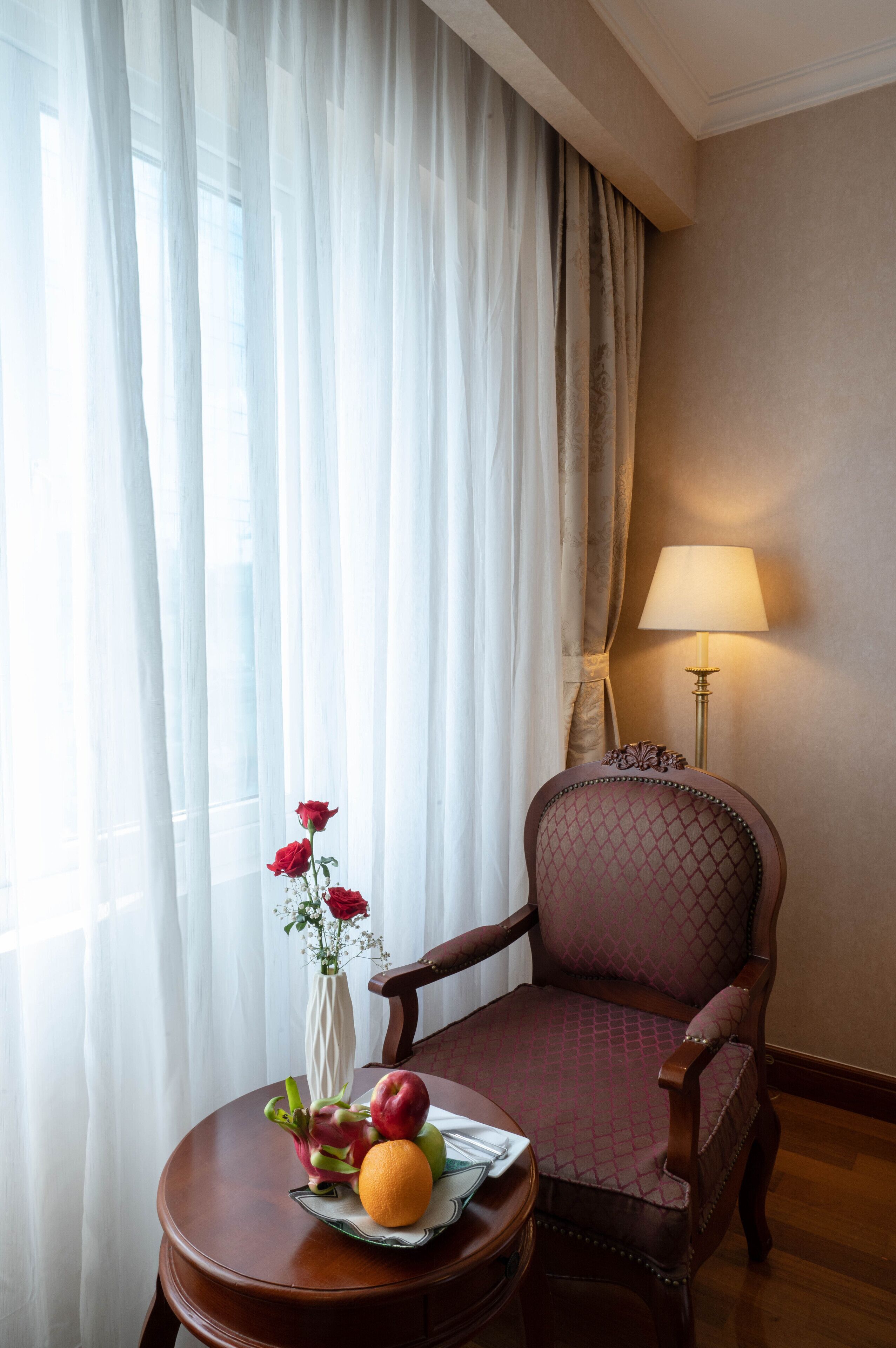 A room with a wooden chair, a round table with fruit and a vase, and a lamp.