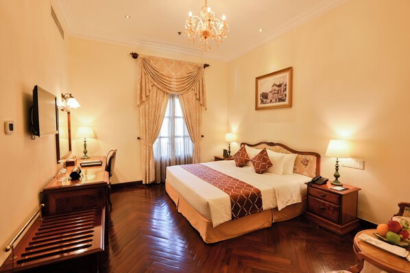 Une chambre d’hôtel avec un grand lit, un plancher en bois, un lustre, des lampes de chevet, un bureau avec une lampe et un tableau au mur.