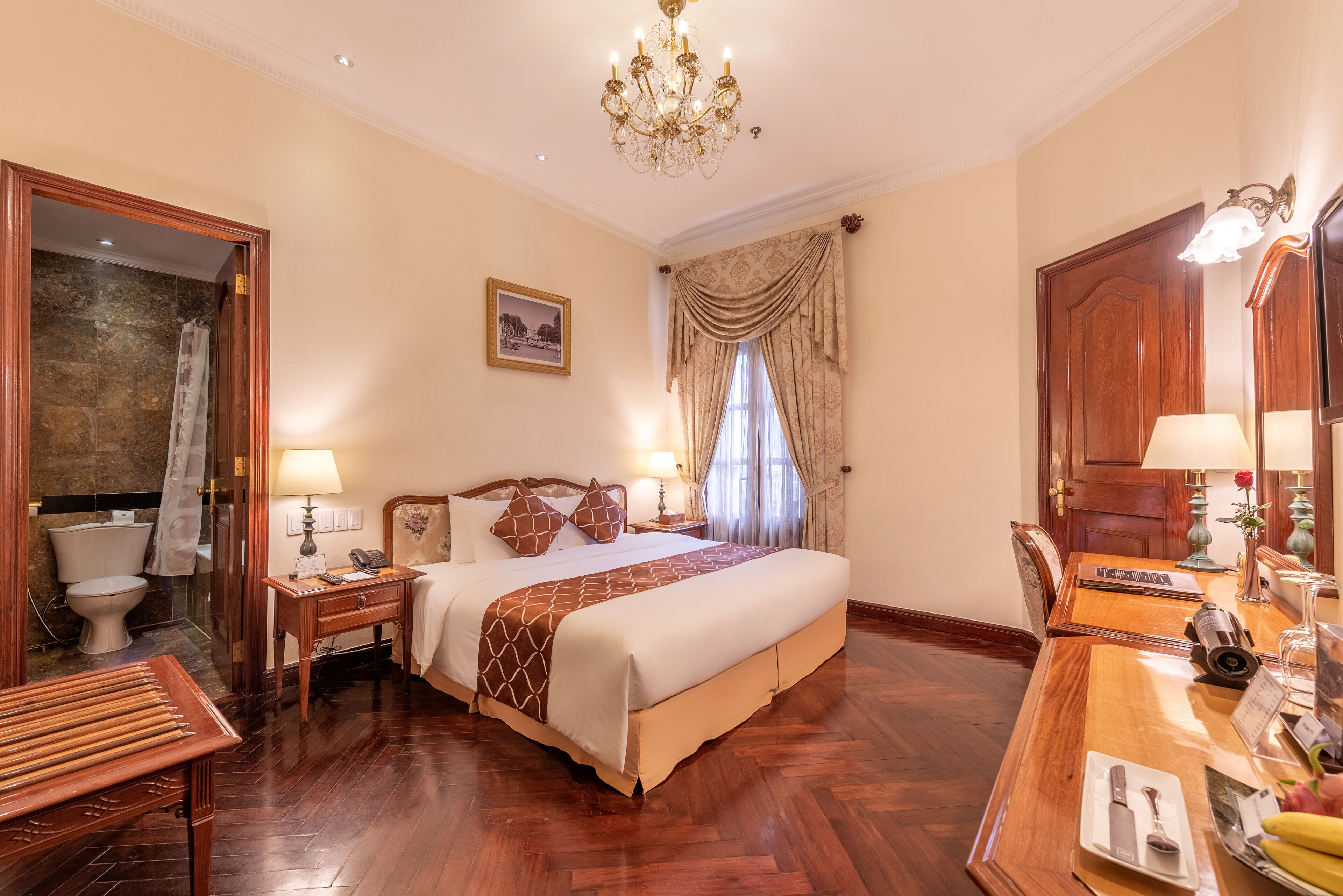 Une chambre d’hôtel avec un grand lit, un plancher en bois, un lustre et un bureau agrémenté d’un miroir et d’une lampe.