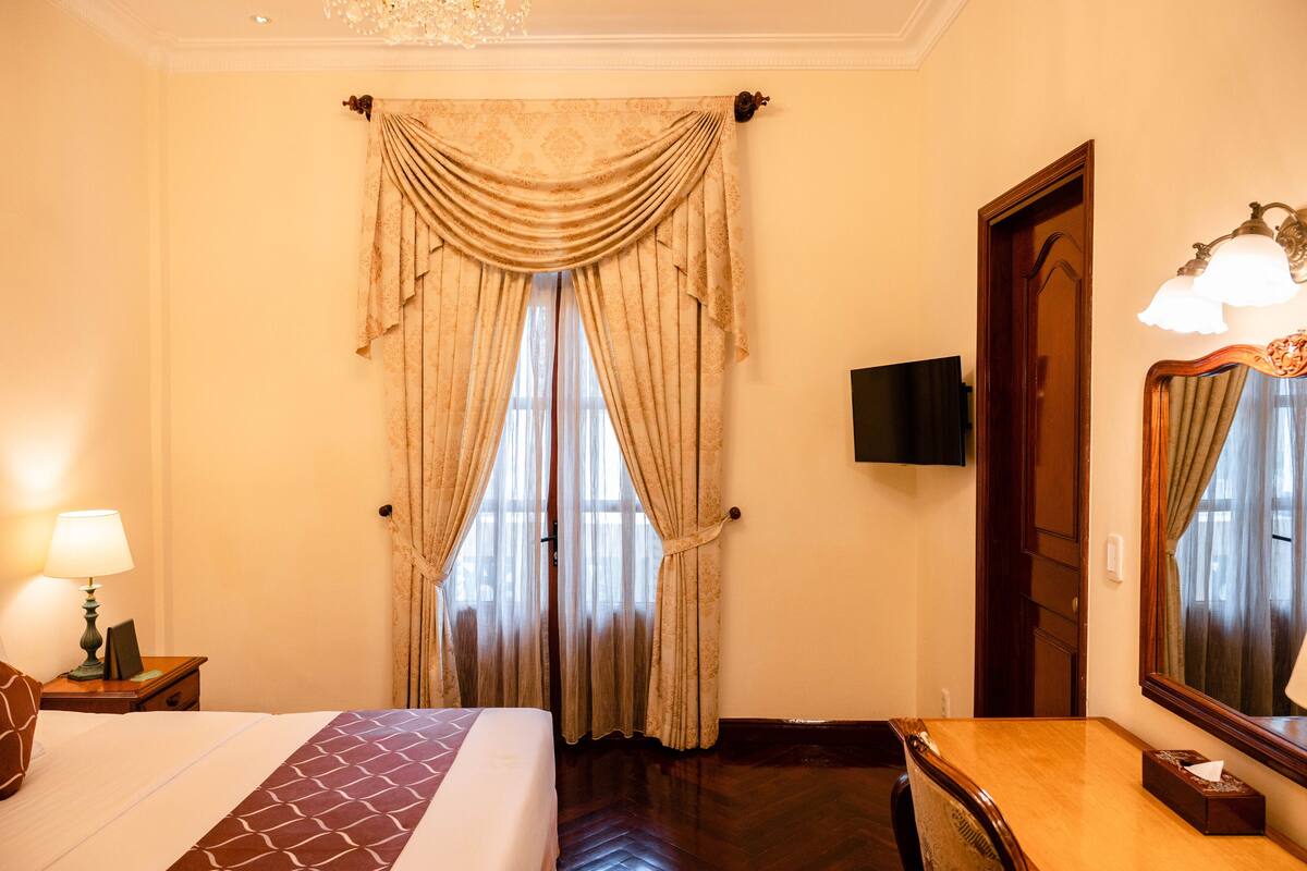A hotel room with a bed, a wooden nightstand with a lamp, a desk with a mirror, and a large window with curtains.