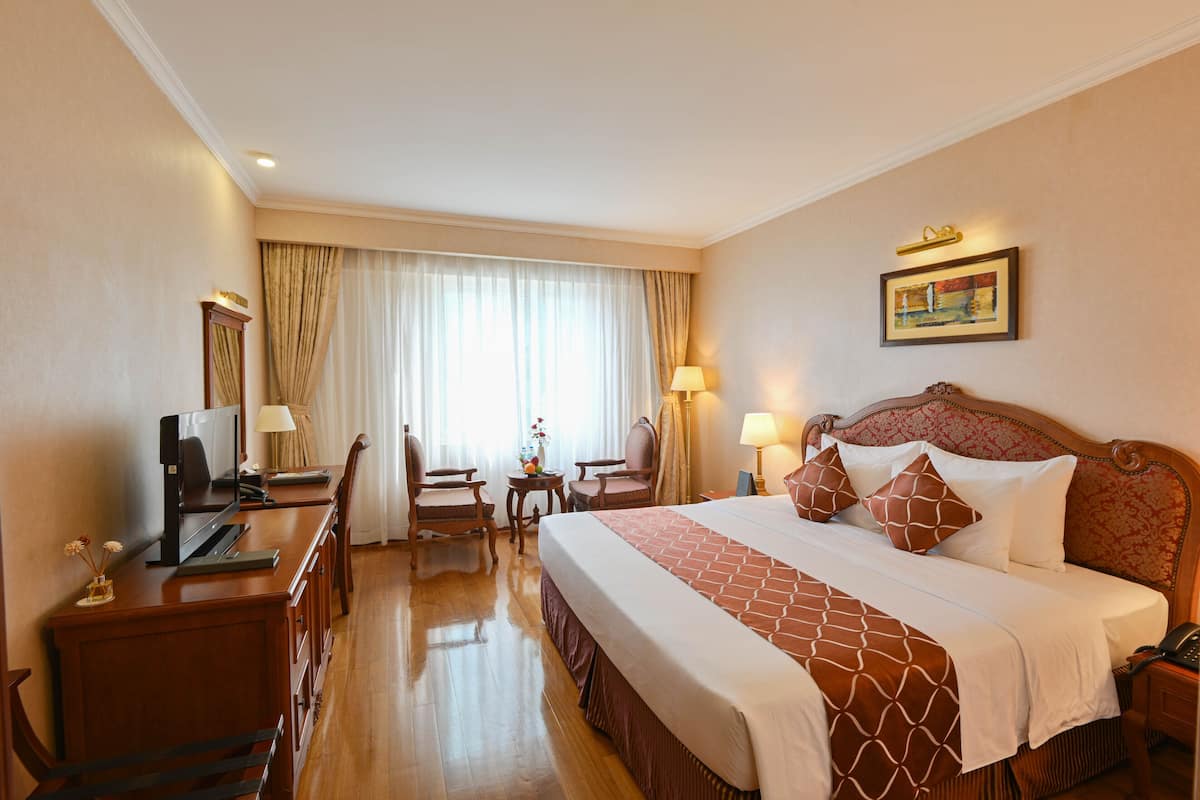A hotel room with a large bed, a desk with a computer, a chair, a small table with a lamp, and a window with curtains.