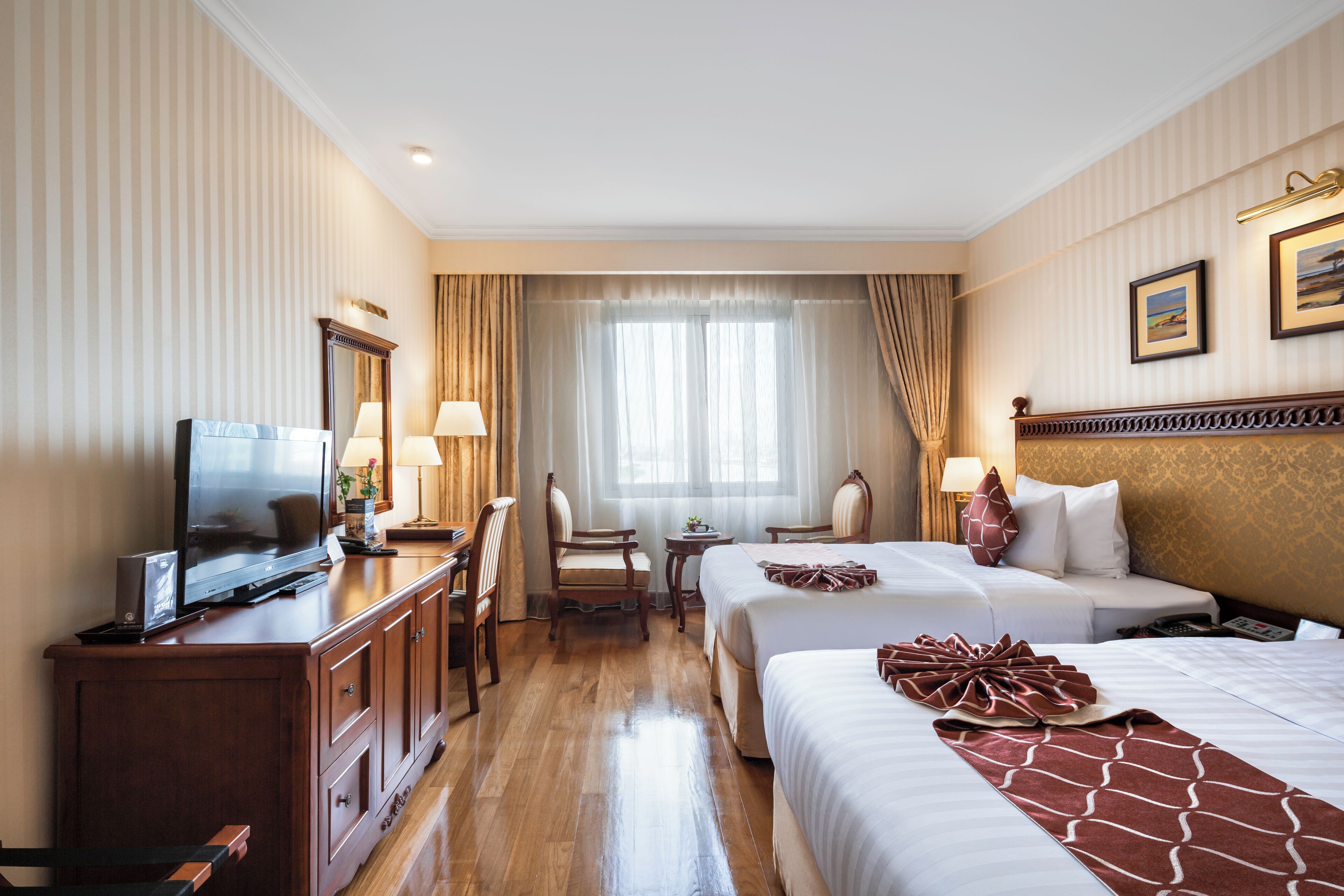 A hotel room with two beds, a TV, a desk with a lamp, and a window with curtains.