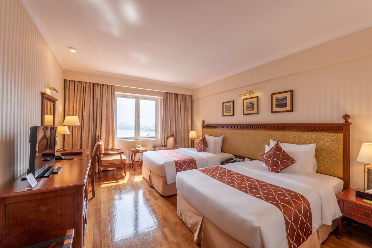 A hotel room with two beds, a desk with a computer, a chair, a small table, and a window with curtains.