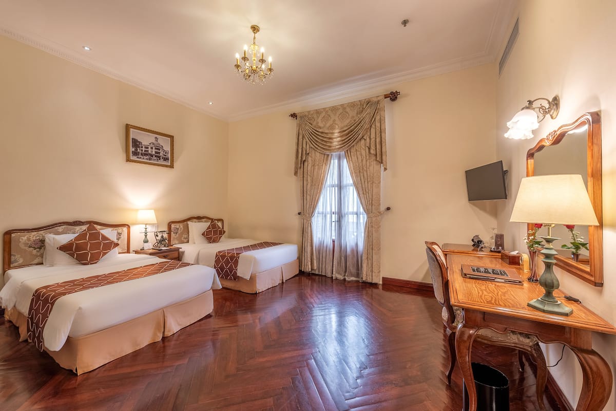 A hotel room with two beds, a desk, a chair, a chandelier, a window with curtains, and a framed picture on the wall.