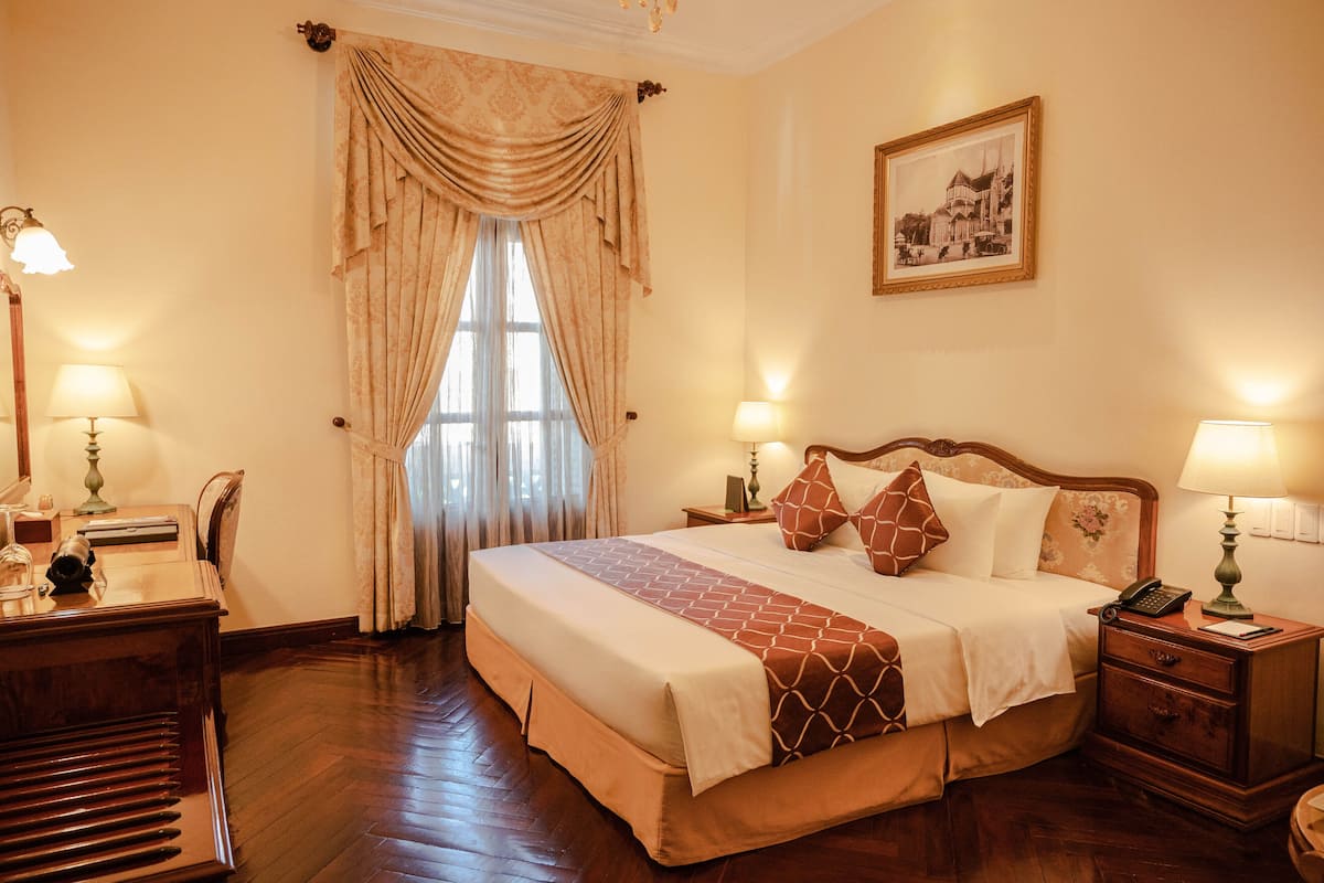 A hotel room with a large bed, two bedside tables with lamps, a desk with a lamp, a chair, and a framed picture on the wall.