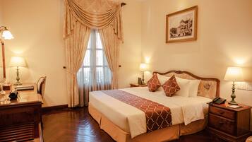 A hotel room with a large bed, two bedside tables with lamps, a desk with a lamp, a chair, and a framed picture on the wall.
