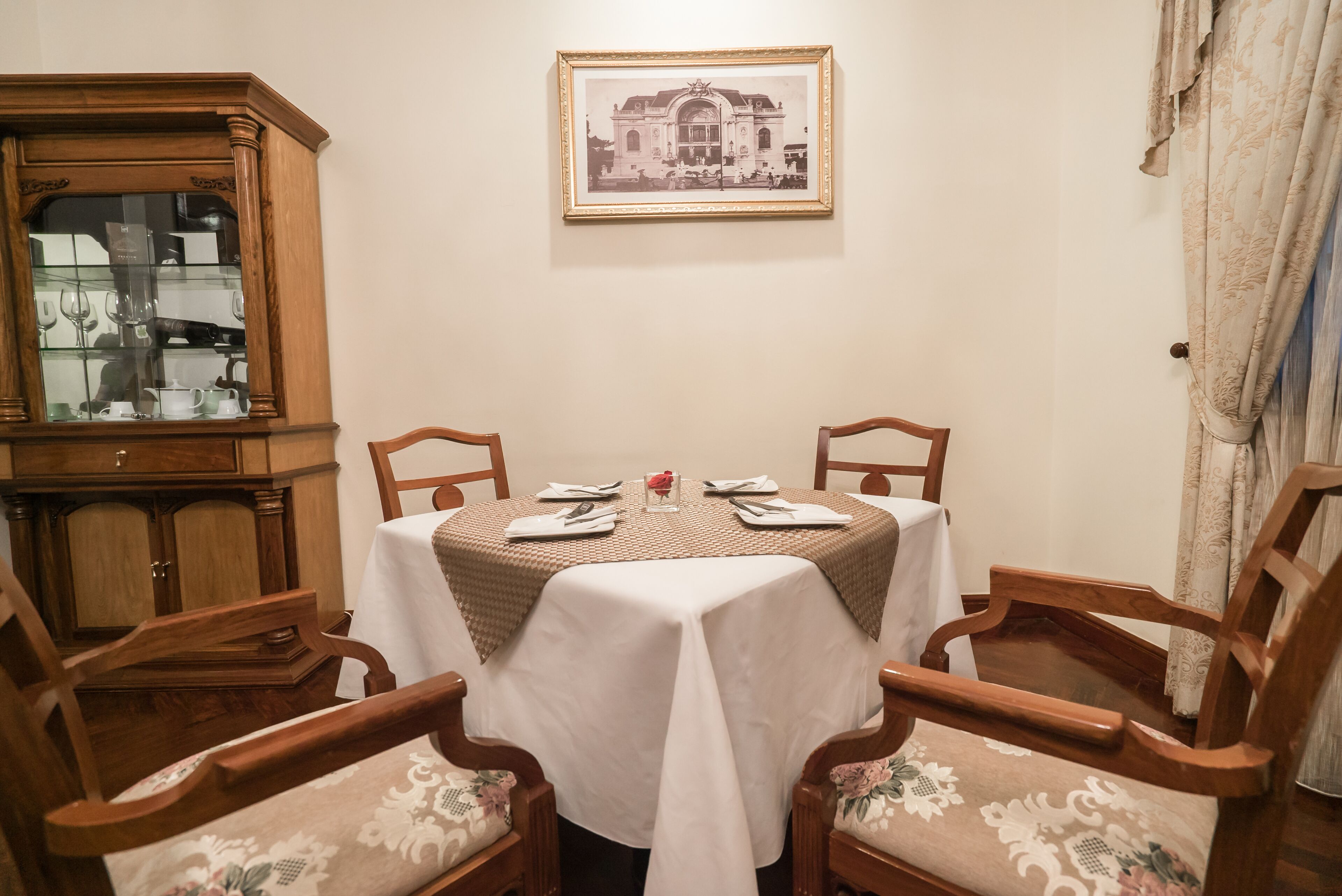 A dining area with a round table set for four, surrounded by wooden chairs with floral upholstery. A wooden cabinet with glass doors is to the left, and a framed historical photograph hangs on the wall above the table.