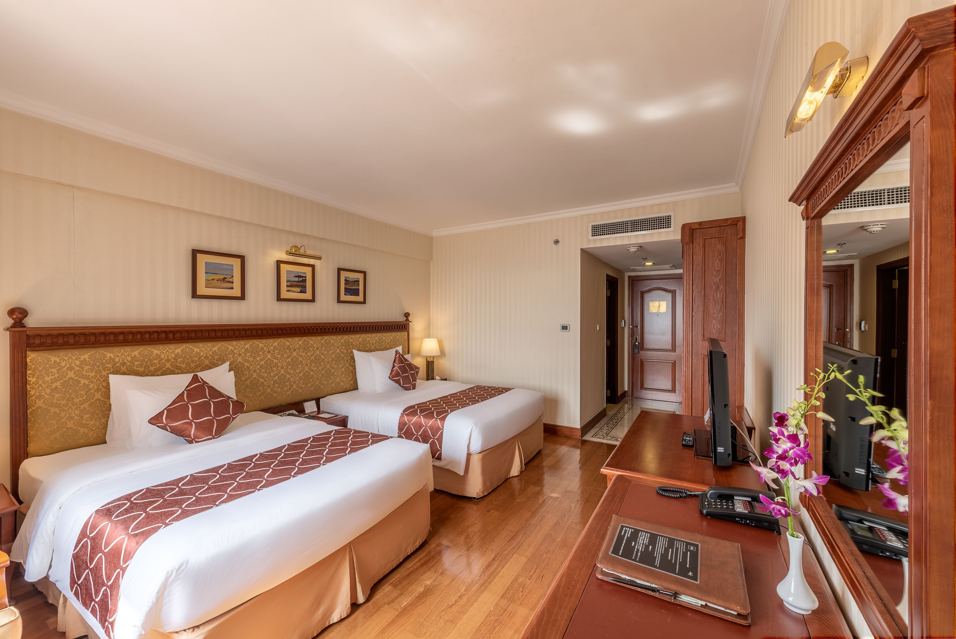 A hotel room with two beds, a desk with a laptop, a vase with flowers, and a mirror.