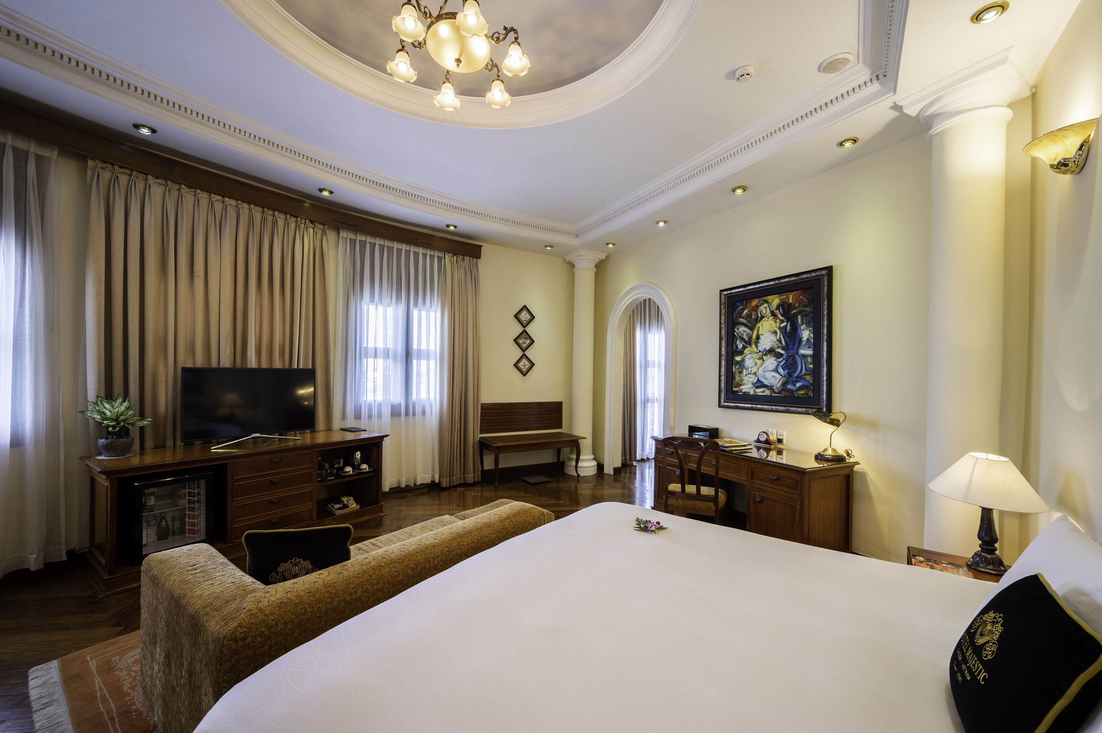Ruime hotelkamer met groot bed, televisie, bureau met lamp en schilderij aan de muur.