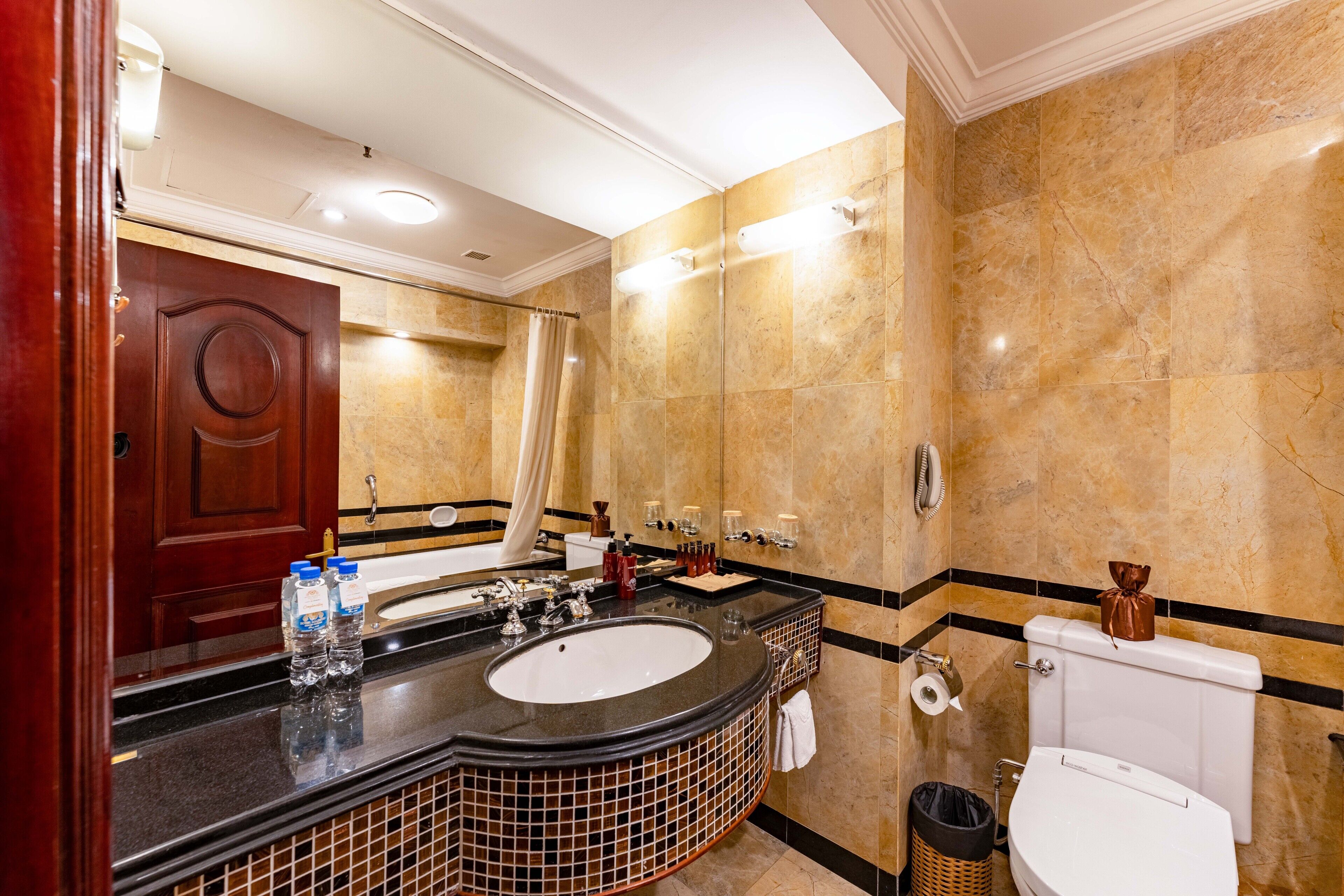 A hotel bathroom with a marble wall, a large sink with a mirror, a toilet, and a shower area.