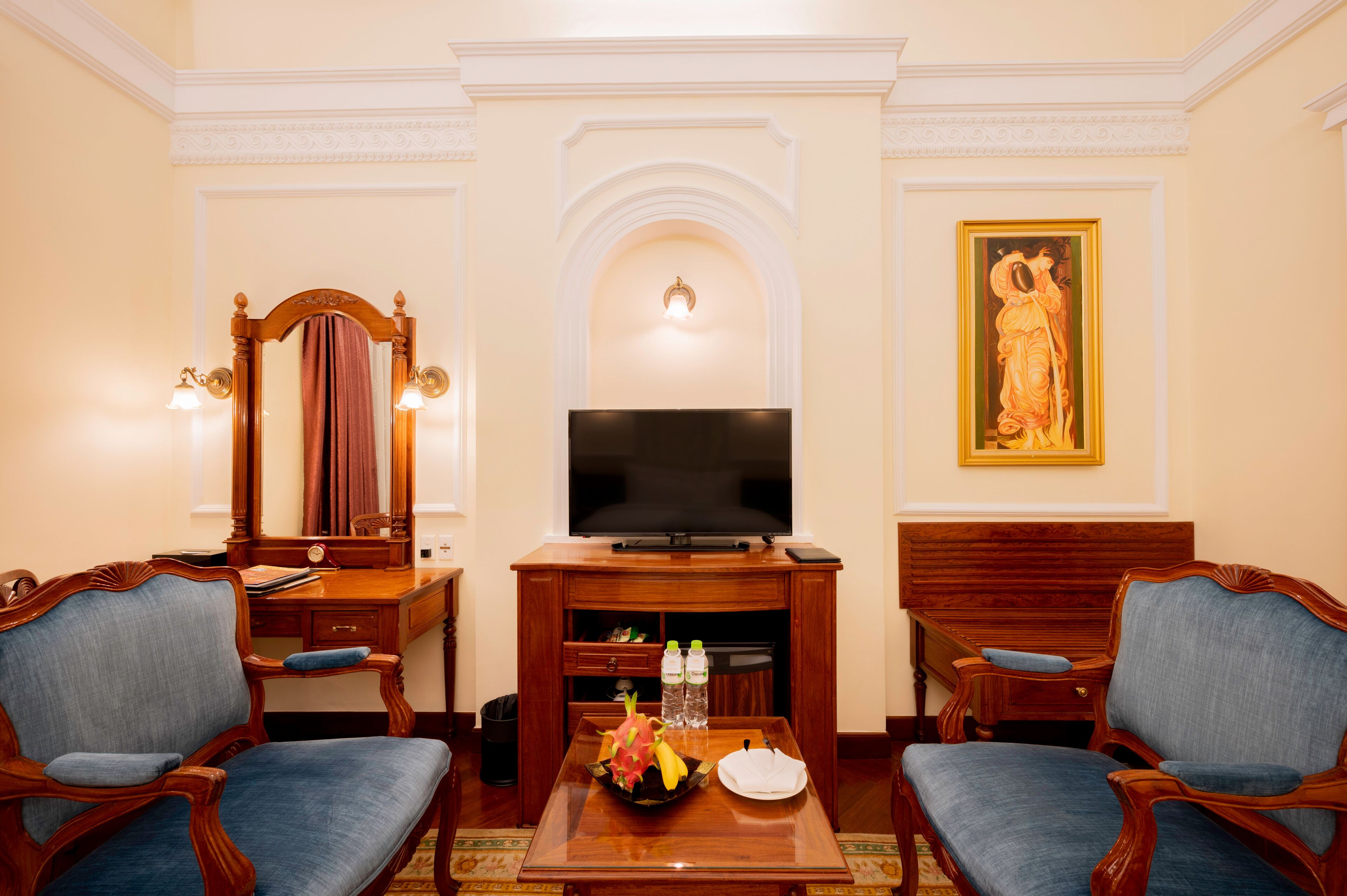 A hotel room with a television, two armchairs, a desk, and a painting on the wall.