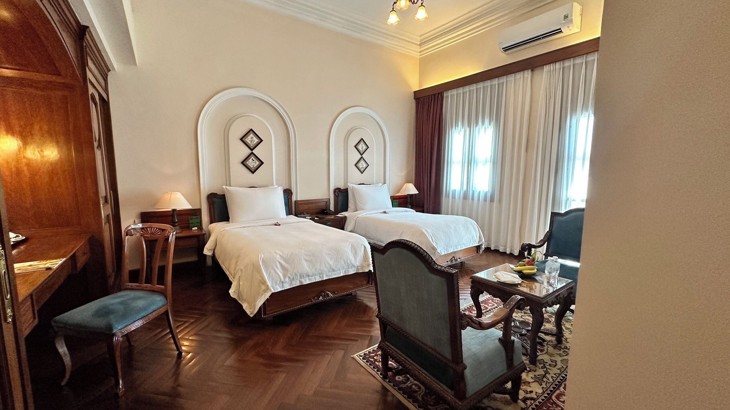 A hotel room with two beds, a desk with a chair, a small table with a lamp, and a window with curtains.
