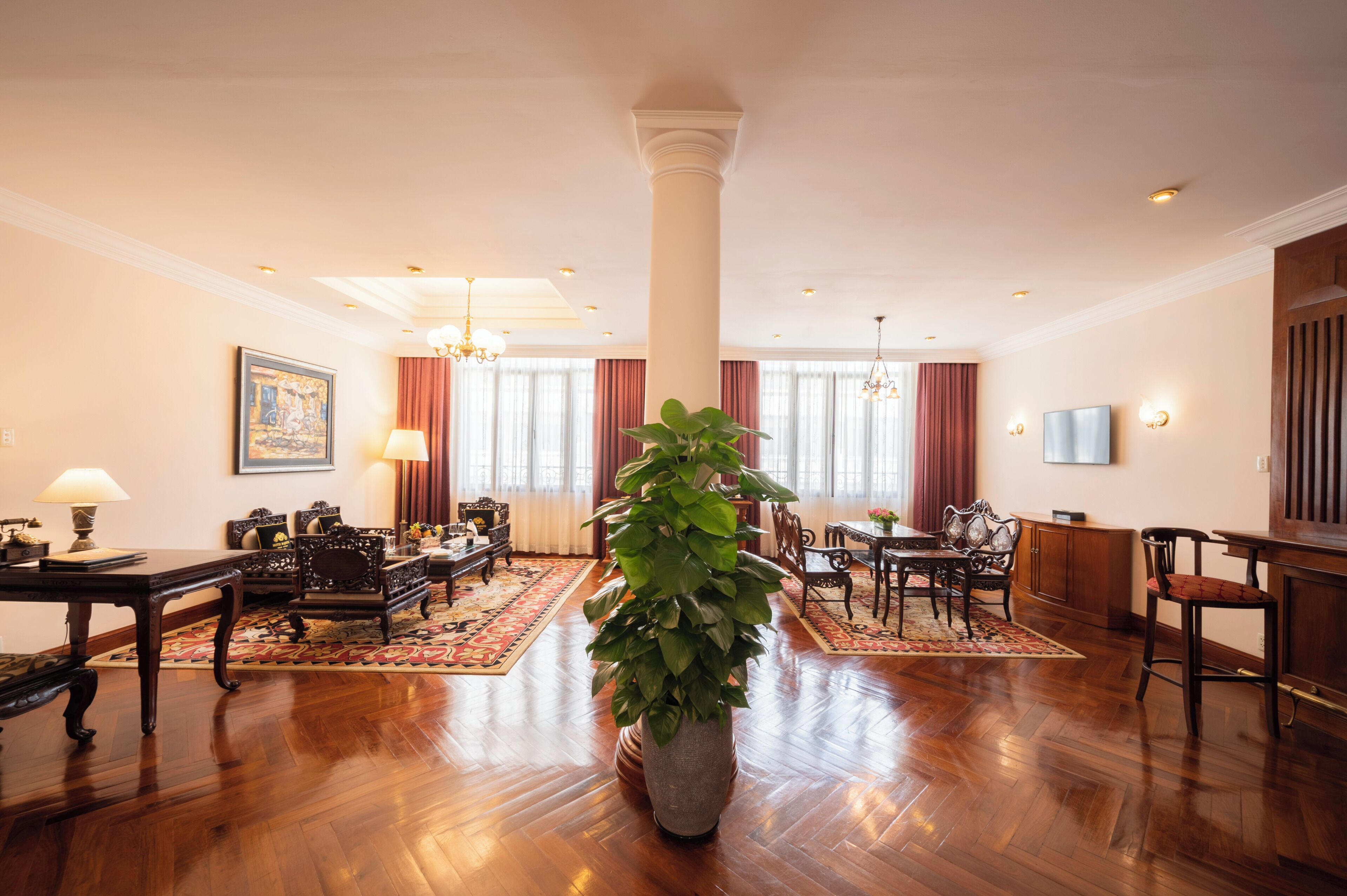 A spacious hotel lobby with a large column, wooden furniture, a central potted plant, and a chandelier.