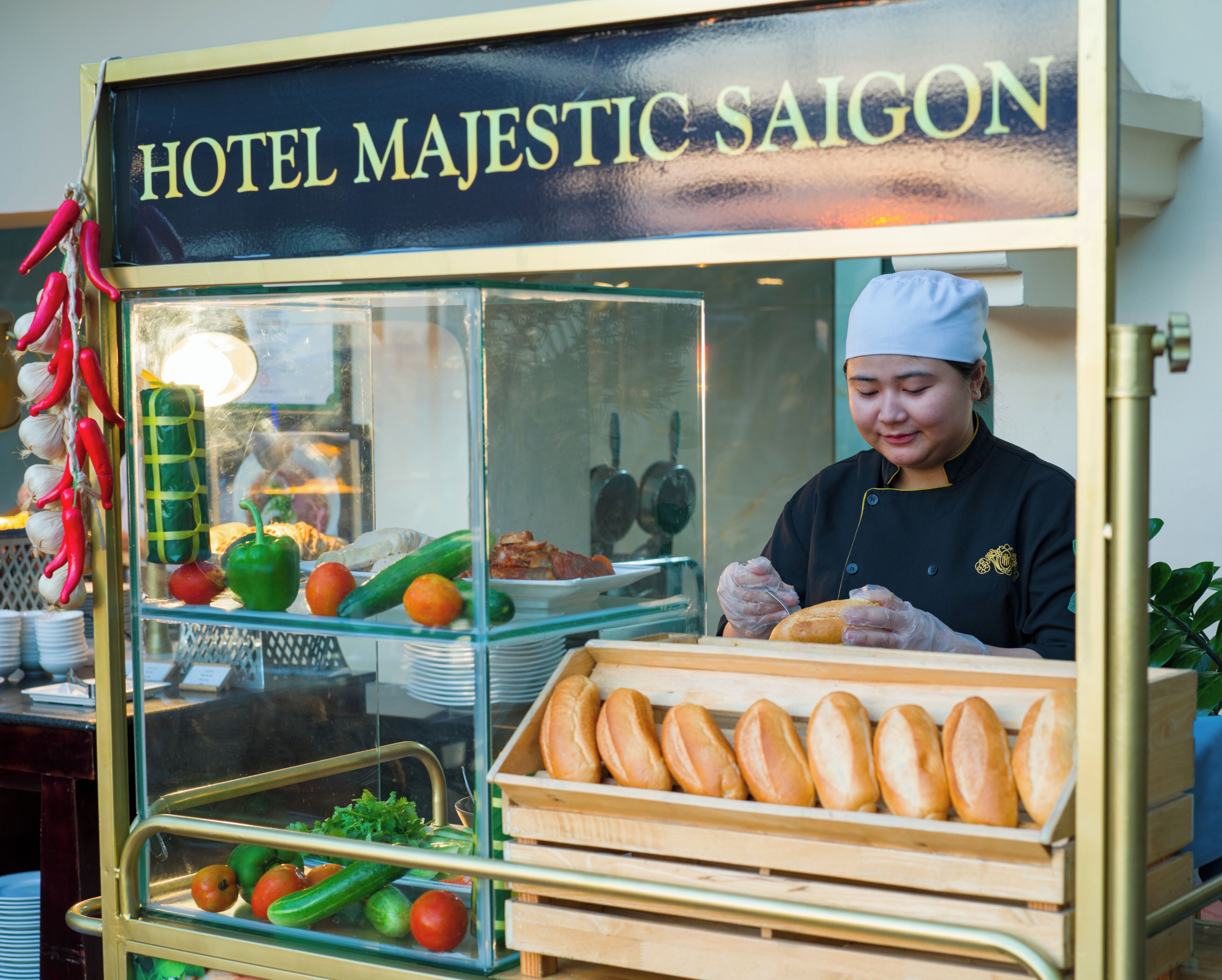 Un carretto di cibo con l'insegna 'HOTEL MAJESTIC SAIGON', uno chef che prepara piatti, un espositore di pane e verdure fresche.