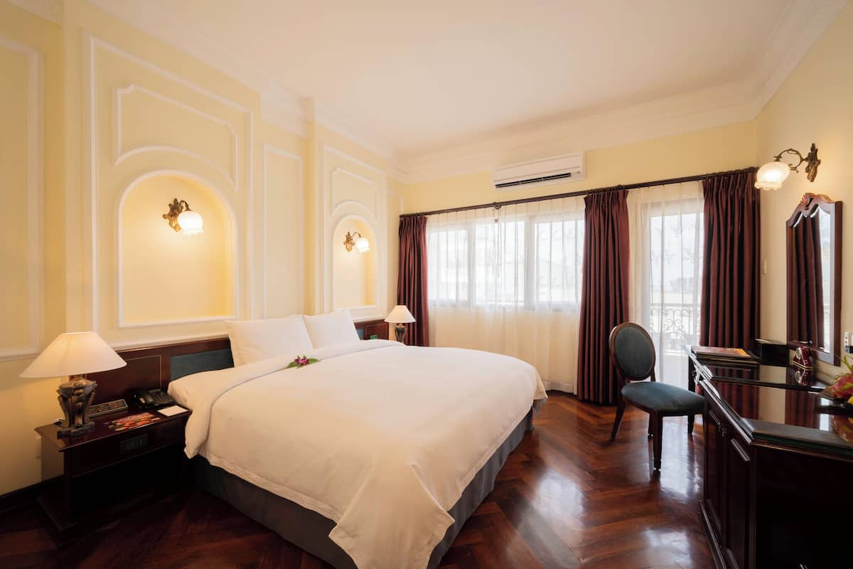 Ein Hotelzimmer mit einem großen Bett, zwei Nachttischen mit Lampen, einem Schreibtisch mit Stuhl und einem großen Fenster mit Vorhängen.