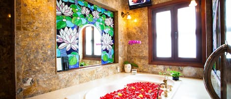 A bathroom with a large bathtub filled with rose petals, a decorative mirror, and a television mounted on the wall.