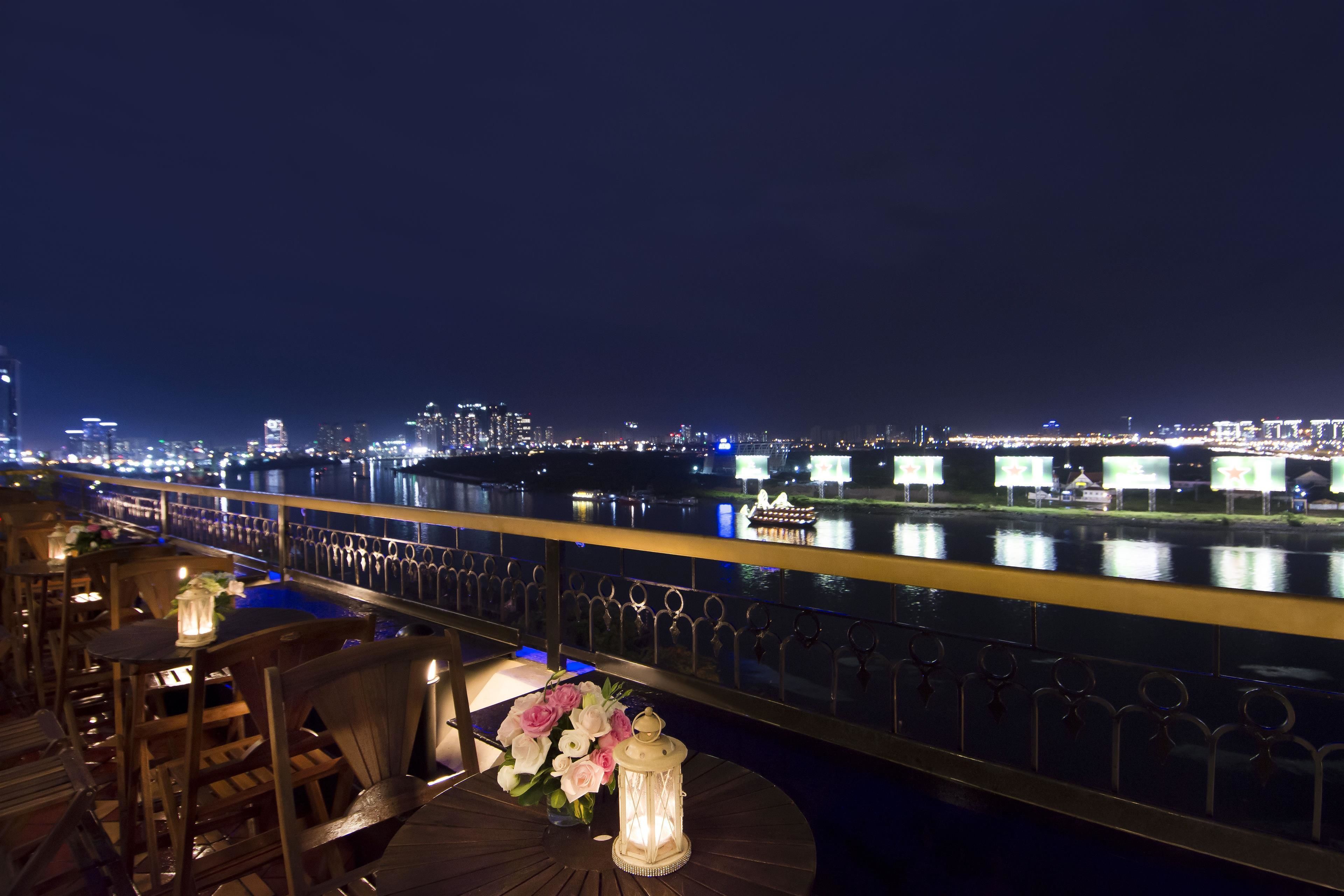 A nighttime view from a balcony with a railing, overlooking a river with city lights in the distance.