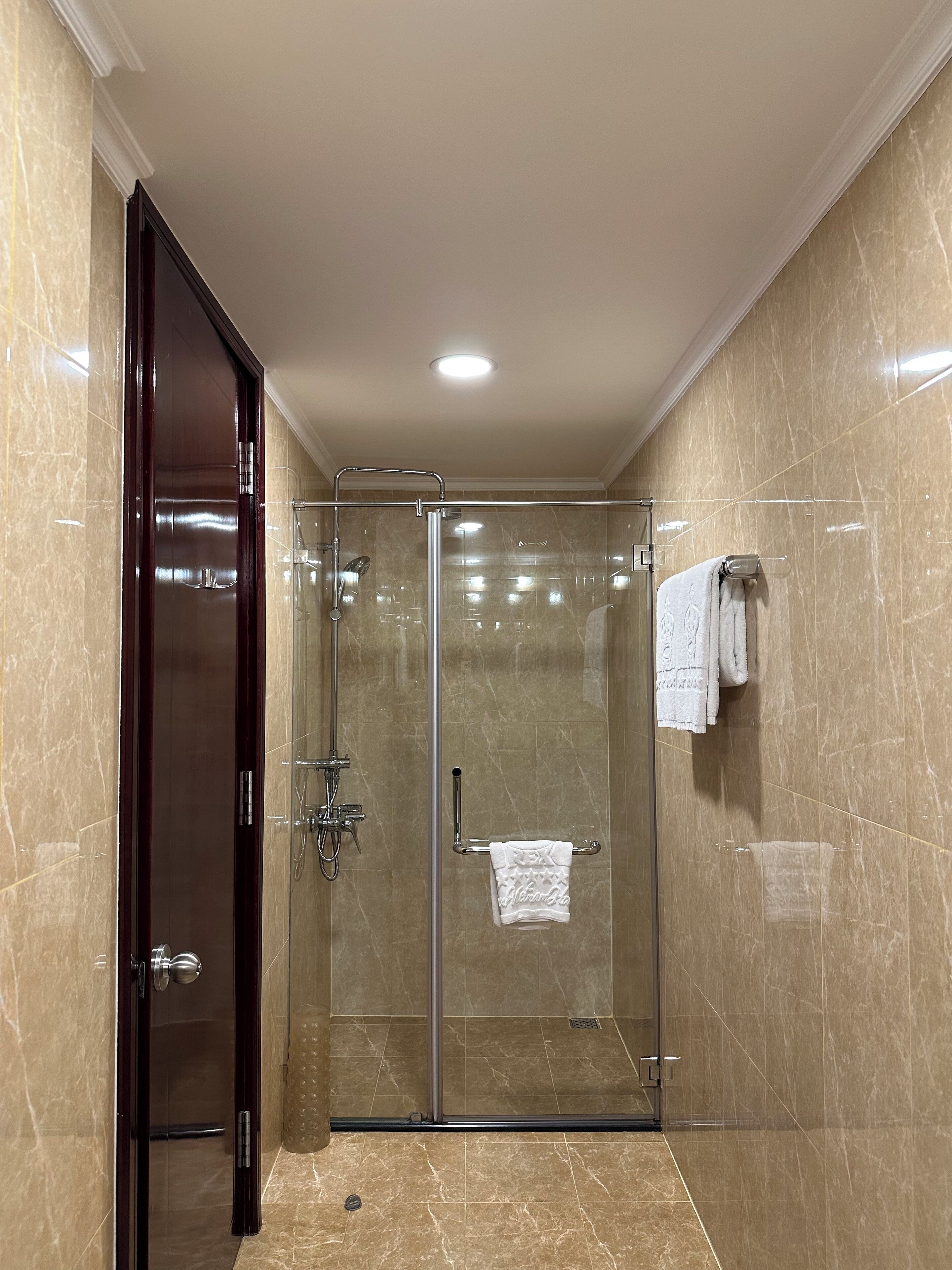 A glass-enclosed shower with a dark wooden door, marble walls, and a towel rack with two towels.