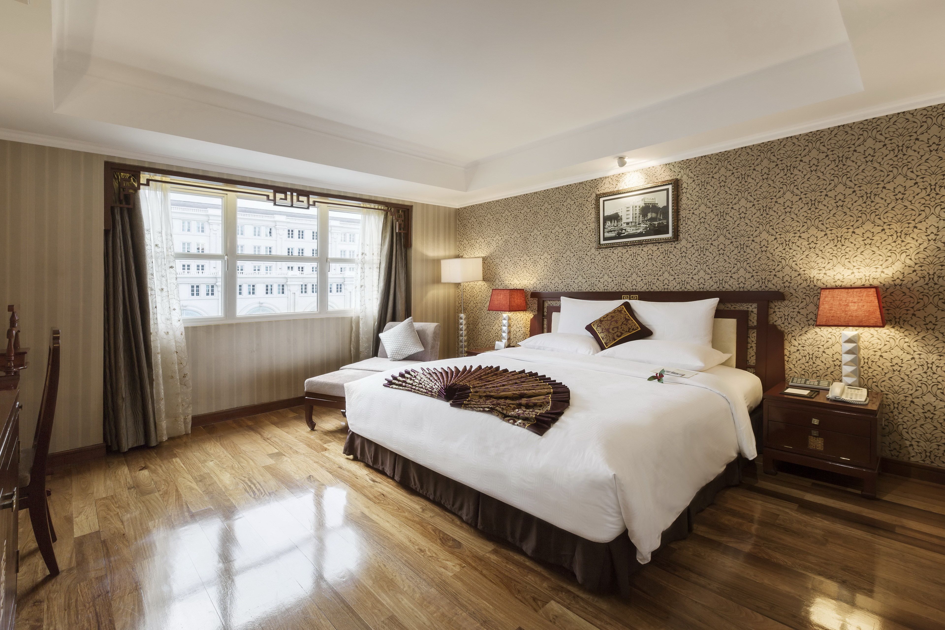 A hotel room with a large bed, two bedside tables, a window with curtains, and a patterned wallpaper.