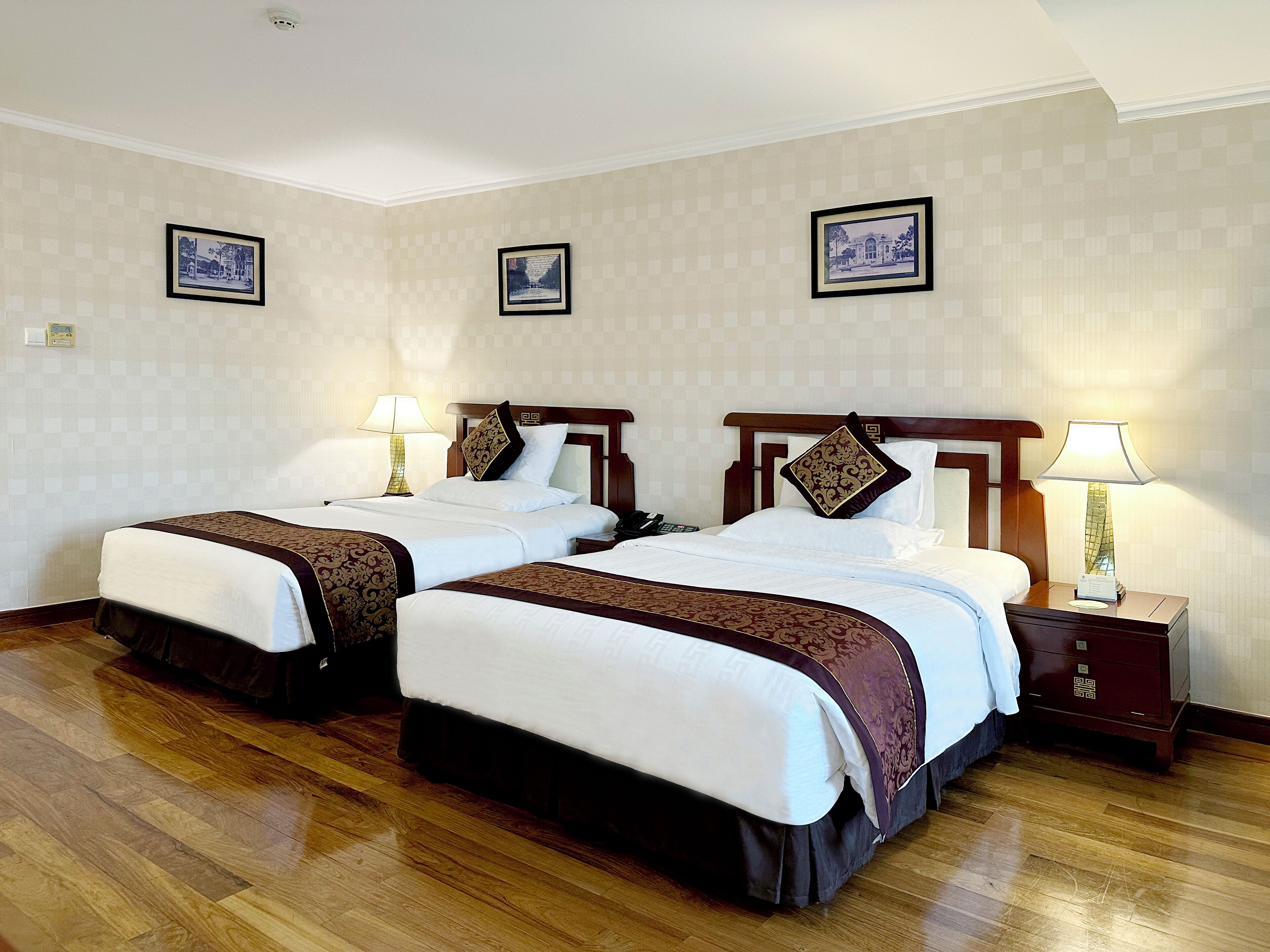 Habitación de hotel con dos camas, cada una con cabecero de madera, ropa de cama blanca y almohadas decorativas. Hay mesitas de noche con lámparas y una caja fuerte pequeña. La habitación tiene un estampado de papel tapiz a cuadros y suelo de madera.