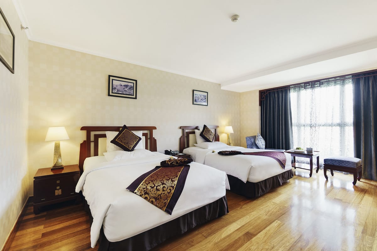 Ein Hotelzimmer mit zwei Betten, Holzboden, einem Nachttisch mit Lampe, einem kleinen Tisch ebenfalls mit Lampe und einem Fenster mit Vorhängen.