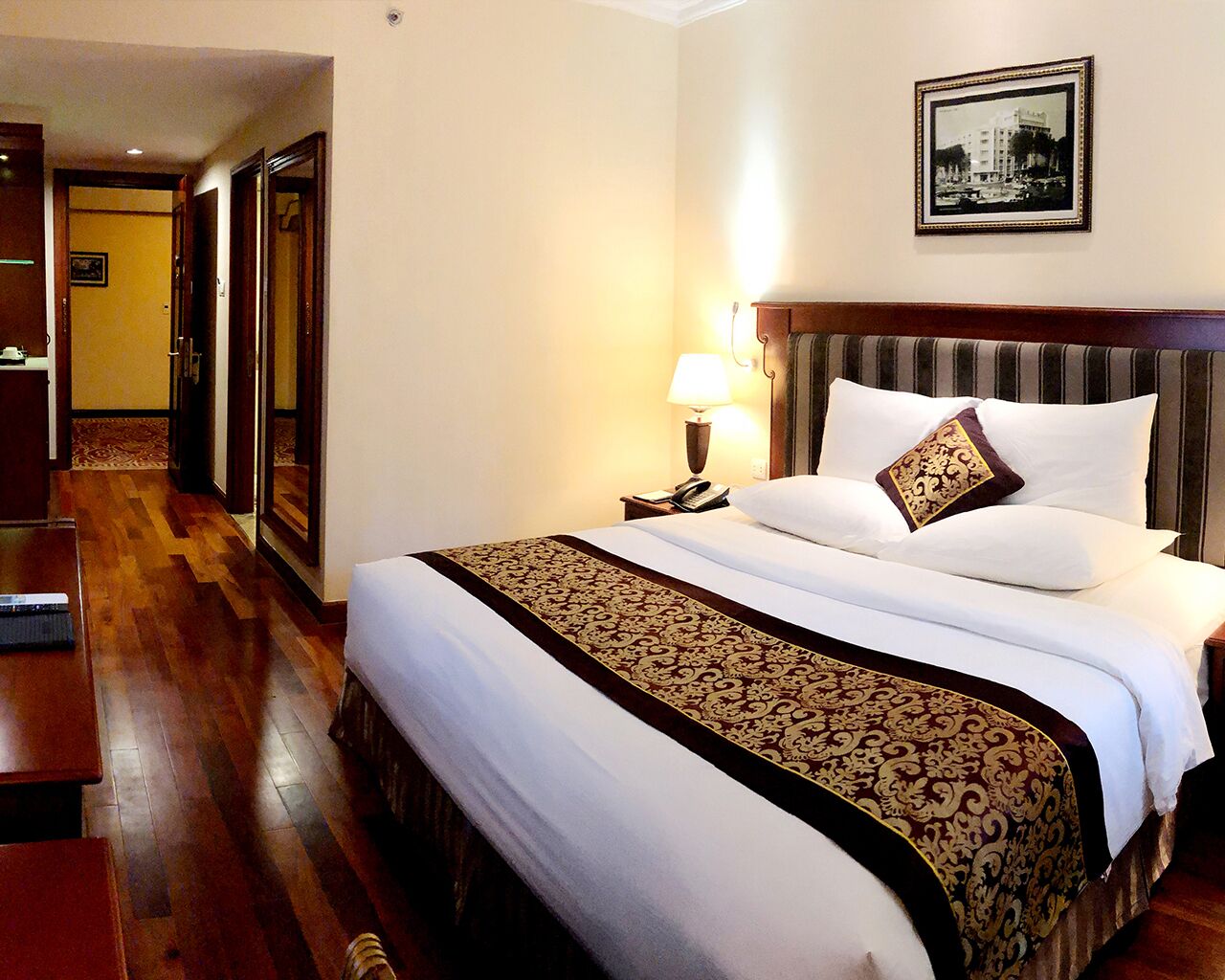 A hotel room with a large bed, a wooden floor, a nightstand with a lamp, and a framed picture on the wall.