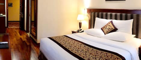A hotel room with a large bed, a wooden floor, a nightstand with a lamp, and a framed picture on the wall.