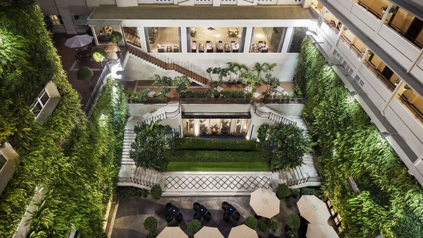Un vestíbulo de hotel de varios niveles con una escalera central, asientos al aire libre y abundante vegetación.
