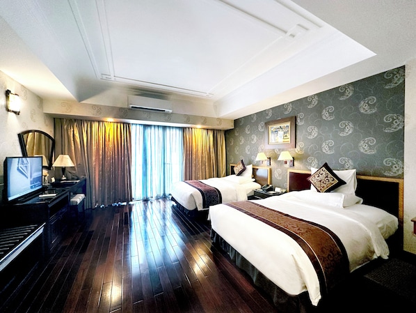 A hotel room with two beds, a TV, a desk, and a large window with curtains.