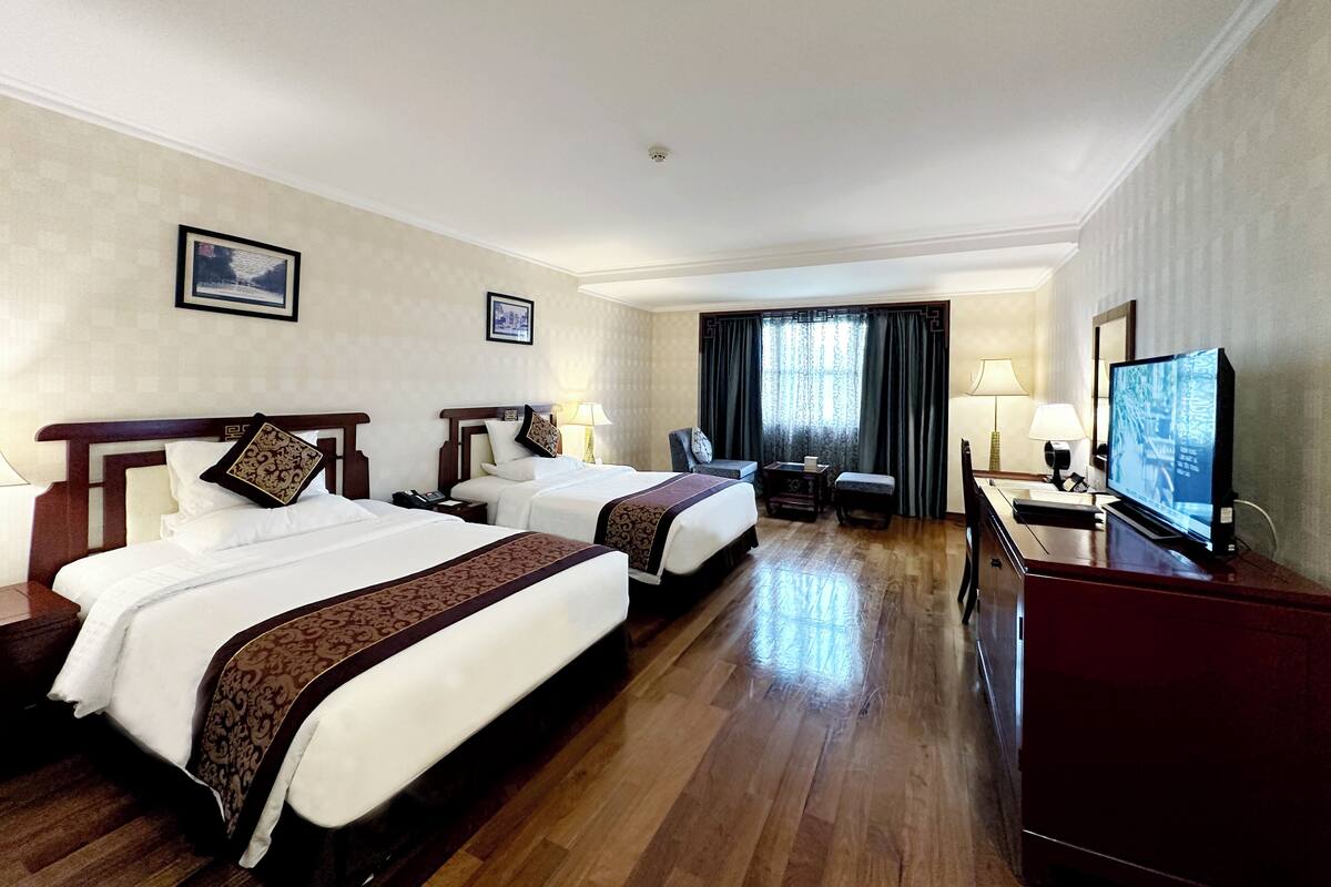 Ein Hotelzimmer mit zwei Betten, einem Schreibtisch, einem Fernseher und einem Fenster mit Vorhängen.