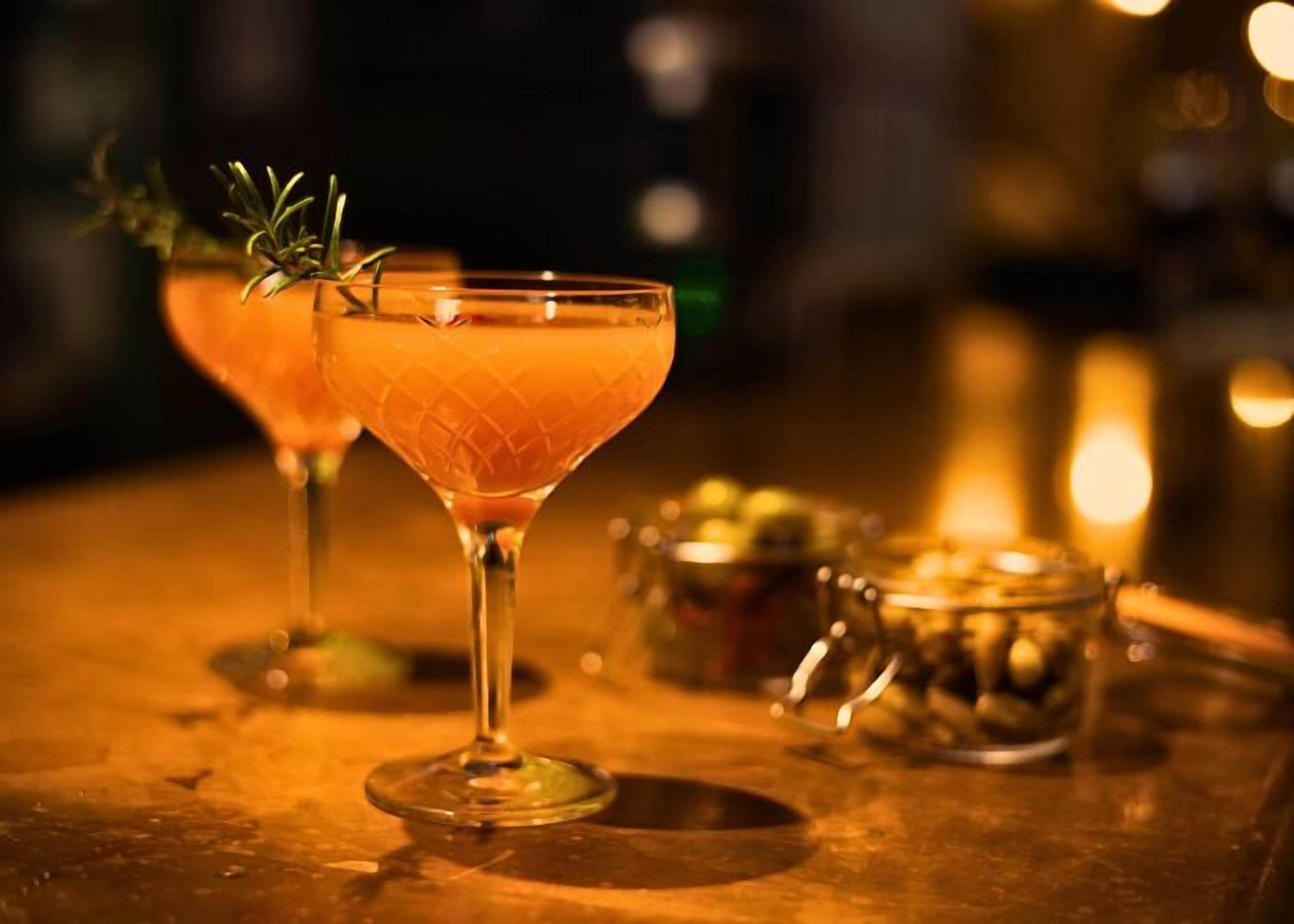 Un bar con dos vasos de cóctel, uno con guarnición, y un recipiente metálico con aceitunas sobre una superficie de madera.