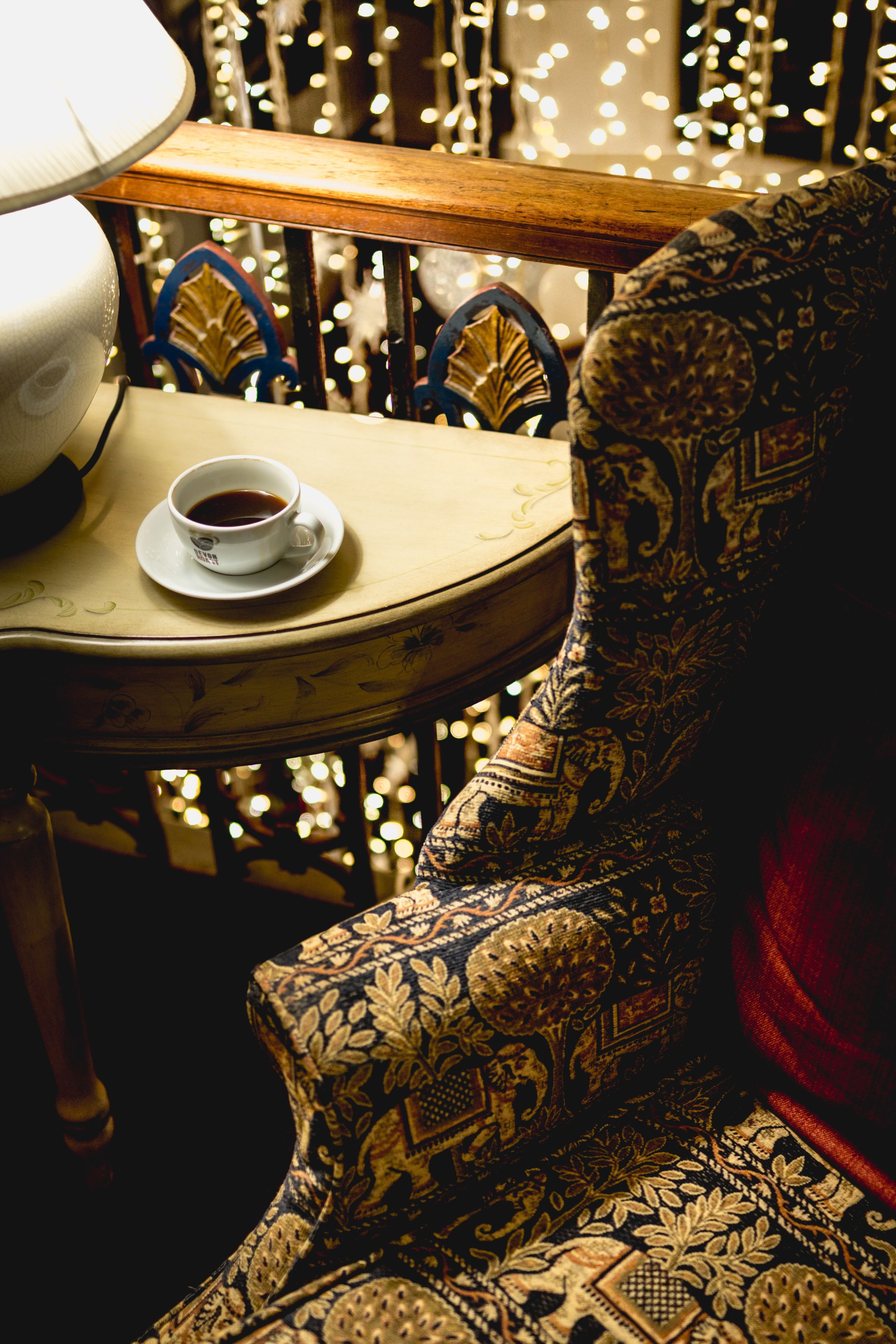 A chair with intricate patterns, a table with a cup of coffee, and a string of lights in the background.