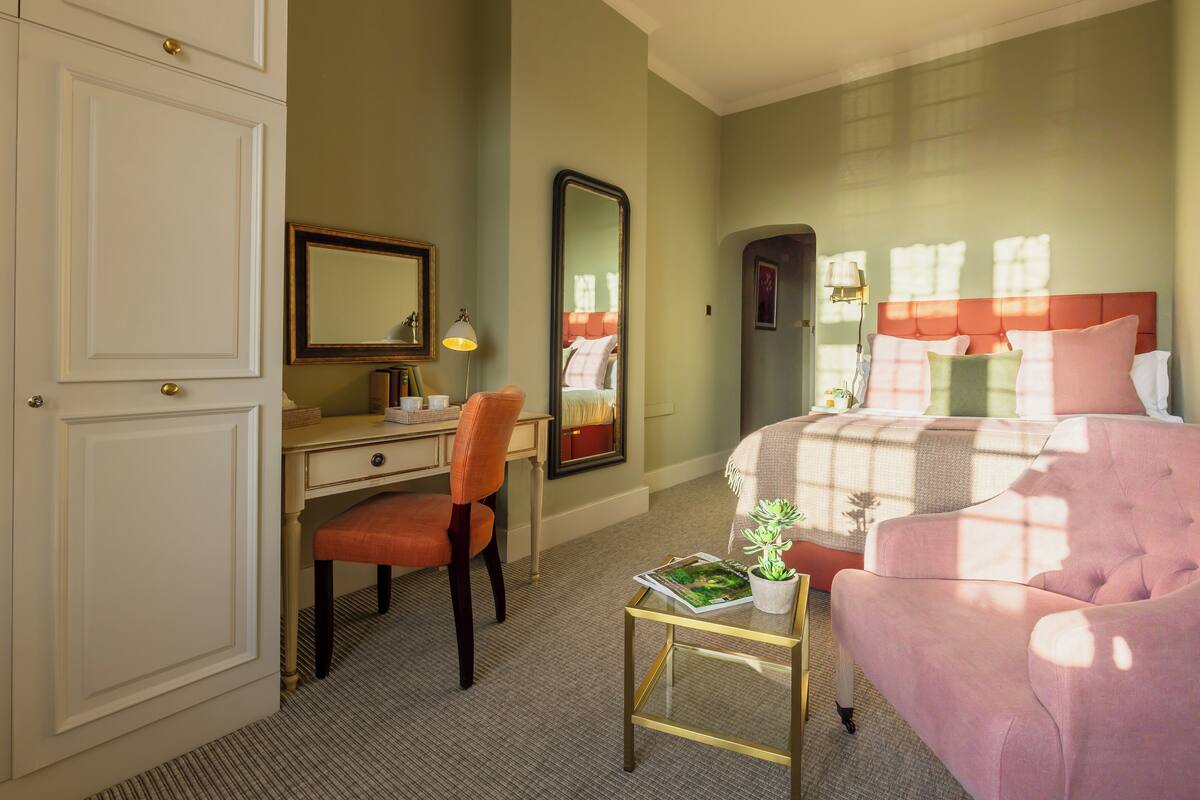 A hotel room with a bed, a desk, a chair, a small table with a plant, and a mirror.