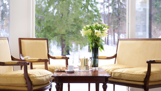 A room with two yellow upholstered chairs, a wooden table with a vase of flowers, a teapot, and a cup. - Vinger Hotell (Kongsvinger)