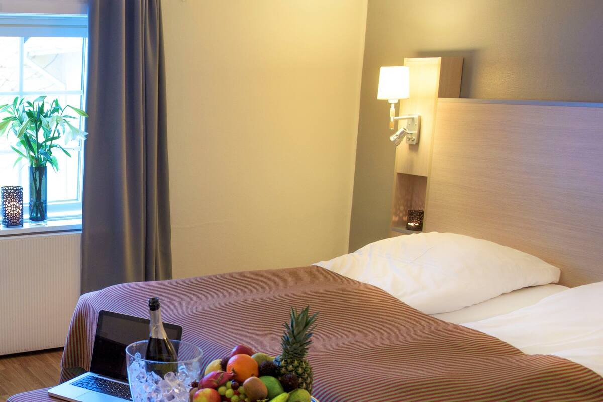 A hotel room with a bed, a laptop on a bedside table, a fruit bowl, and a bottle of champagne.