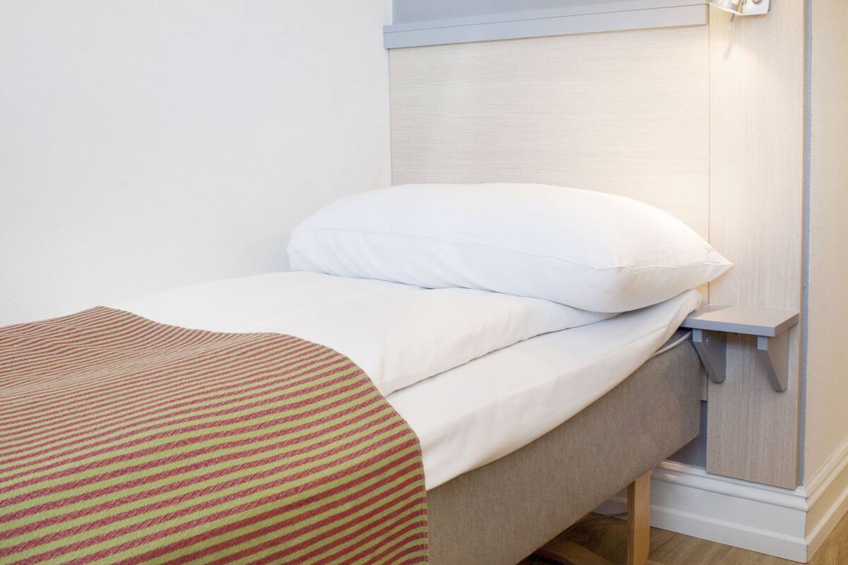 A small, neatly made bed with a white pillow and a striped blanket, a bedside lamp, and a wooden floor.
