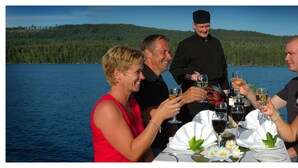 A group of people dining at a table by a lake with a forested hill in the background. - Vinger Hotell (Kongsvinger)