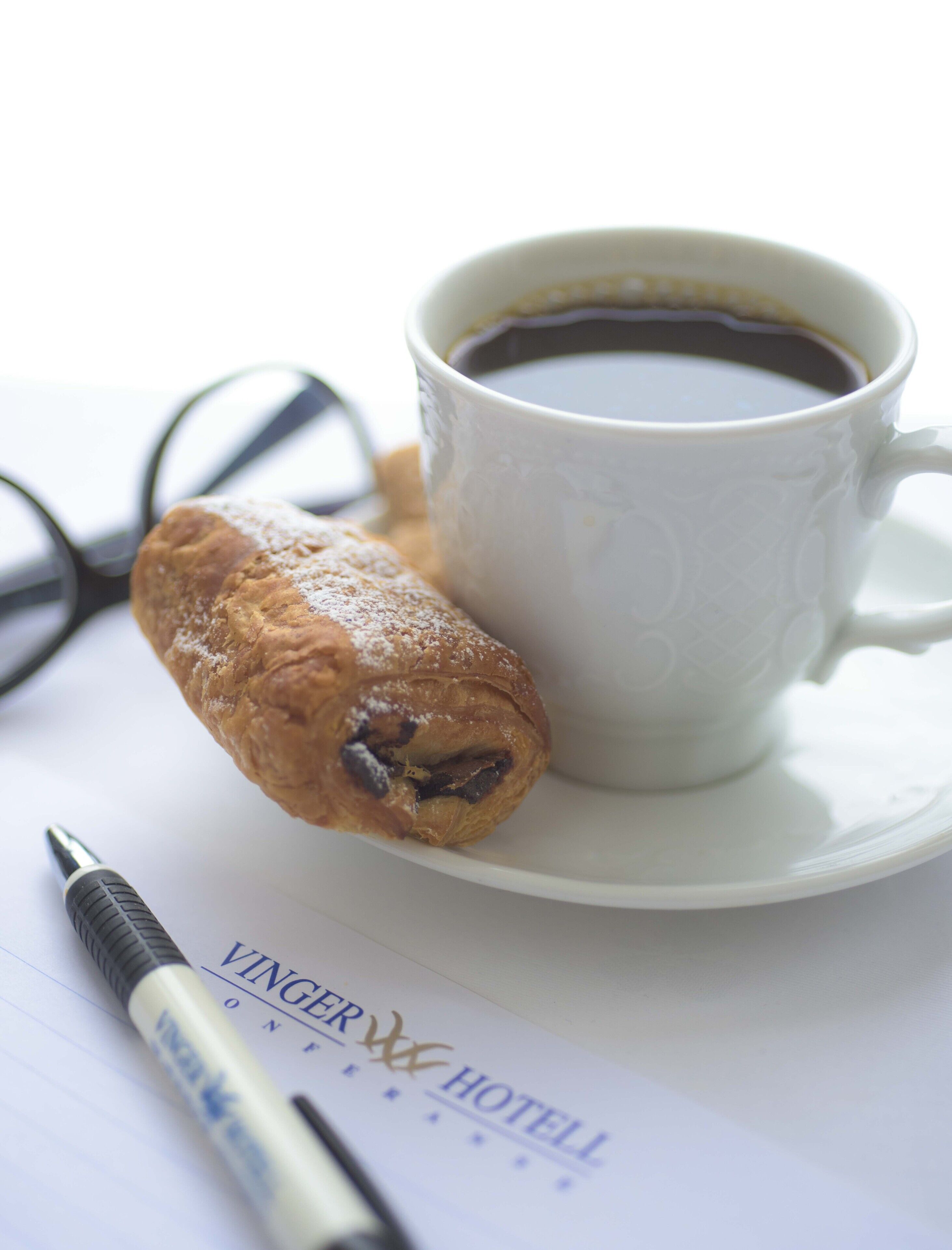 Une tasse de café, une pâtisserie, une paire de lunettes et un stylo sont posés sur une table, et on peut voir le nom de l’hôtel.